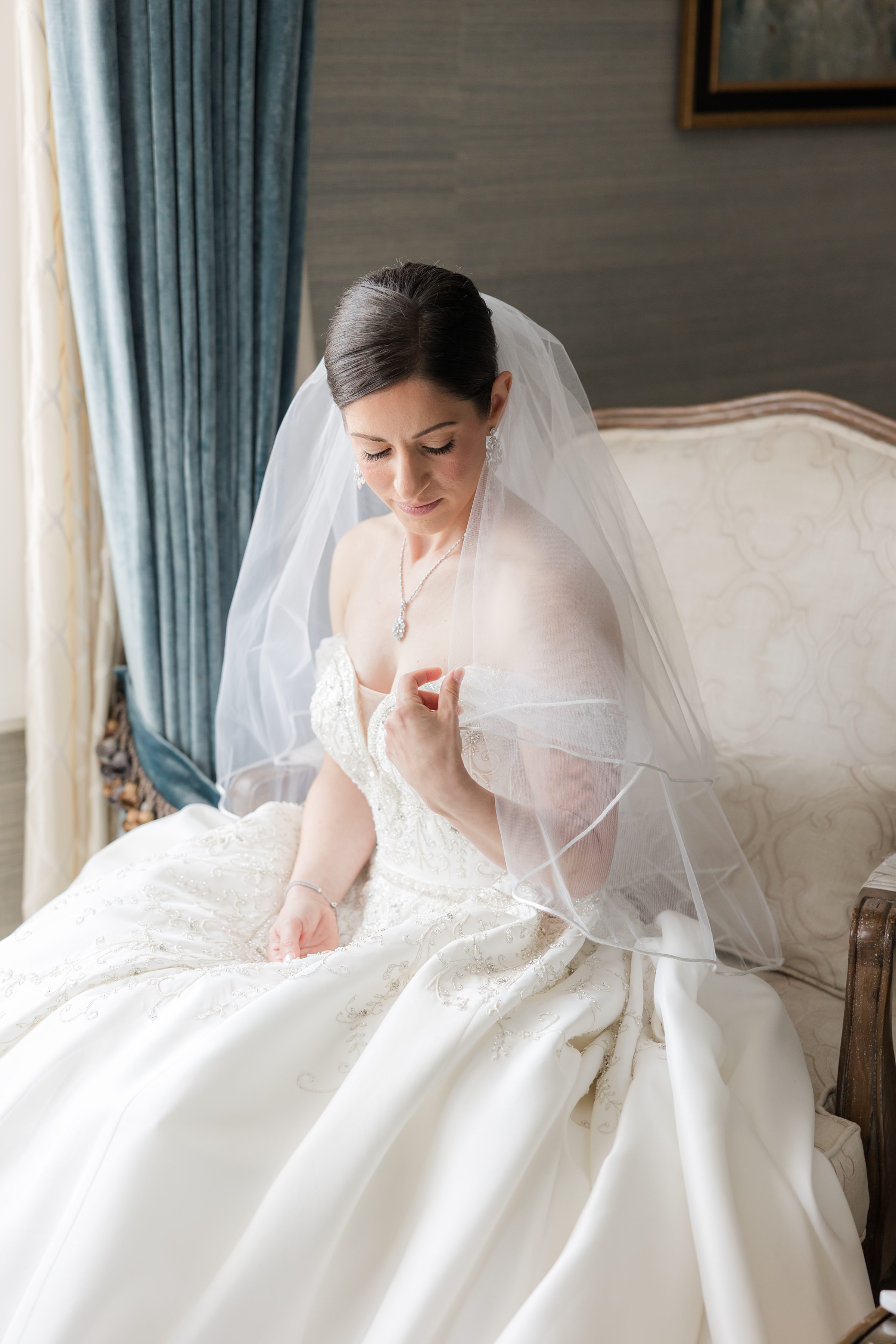 Bride holding her veil and looking at her hand, seated on a wooden chair with white cushion, by the window with white and blue curtains