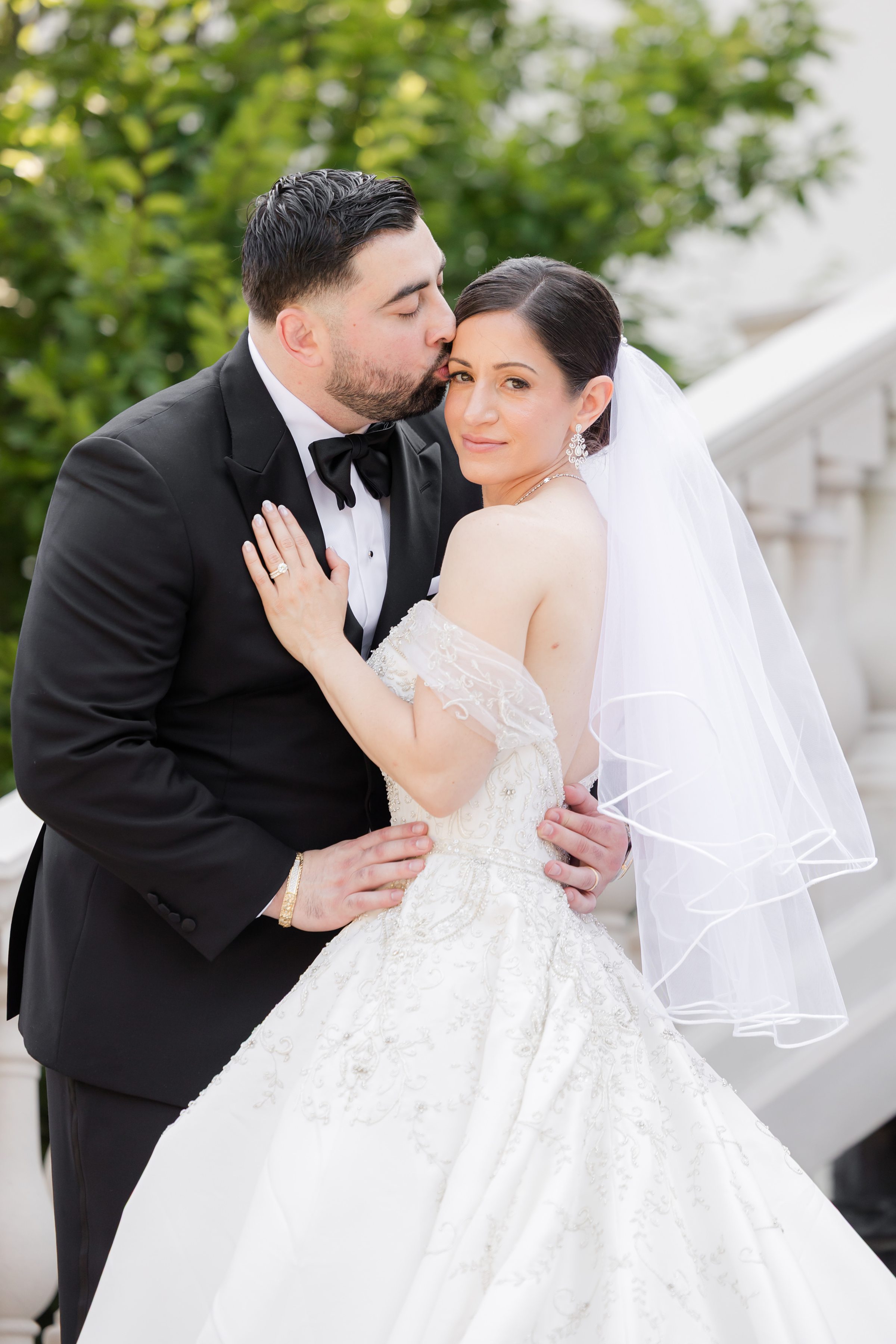 Half-body portrait of groom kissing the bride's forehead while her hand is on his chest, and looking into the camera