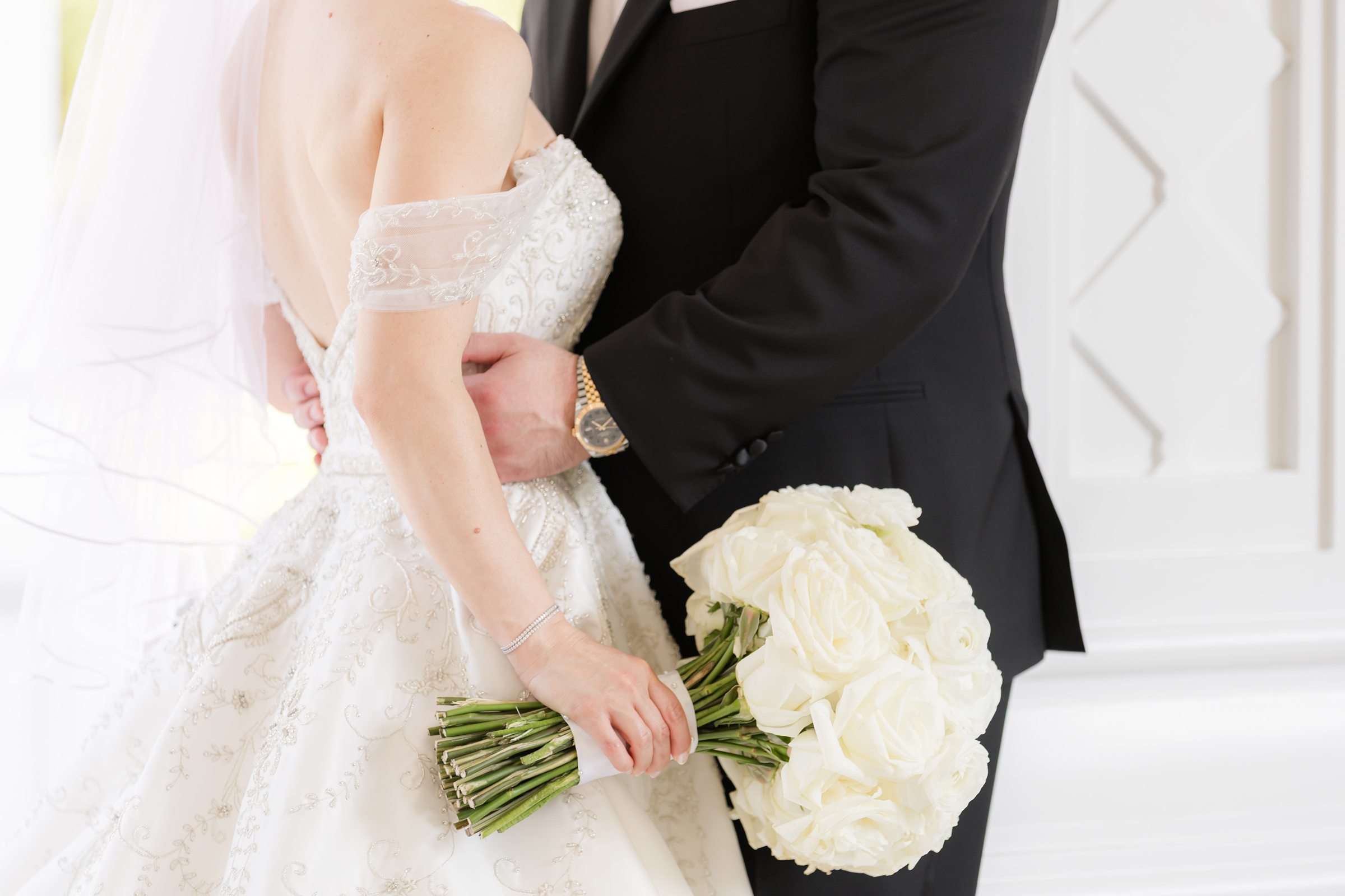 Close-up detail shot of groom holding the bride's waist, and her holding her bouquet with her right hand. Groom's rolex watch is peeking through his tux