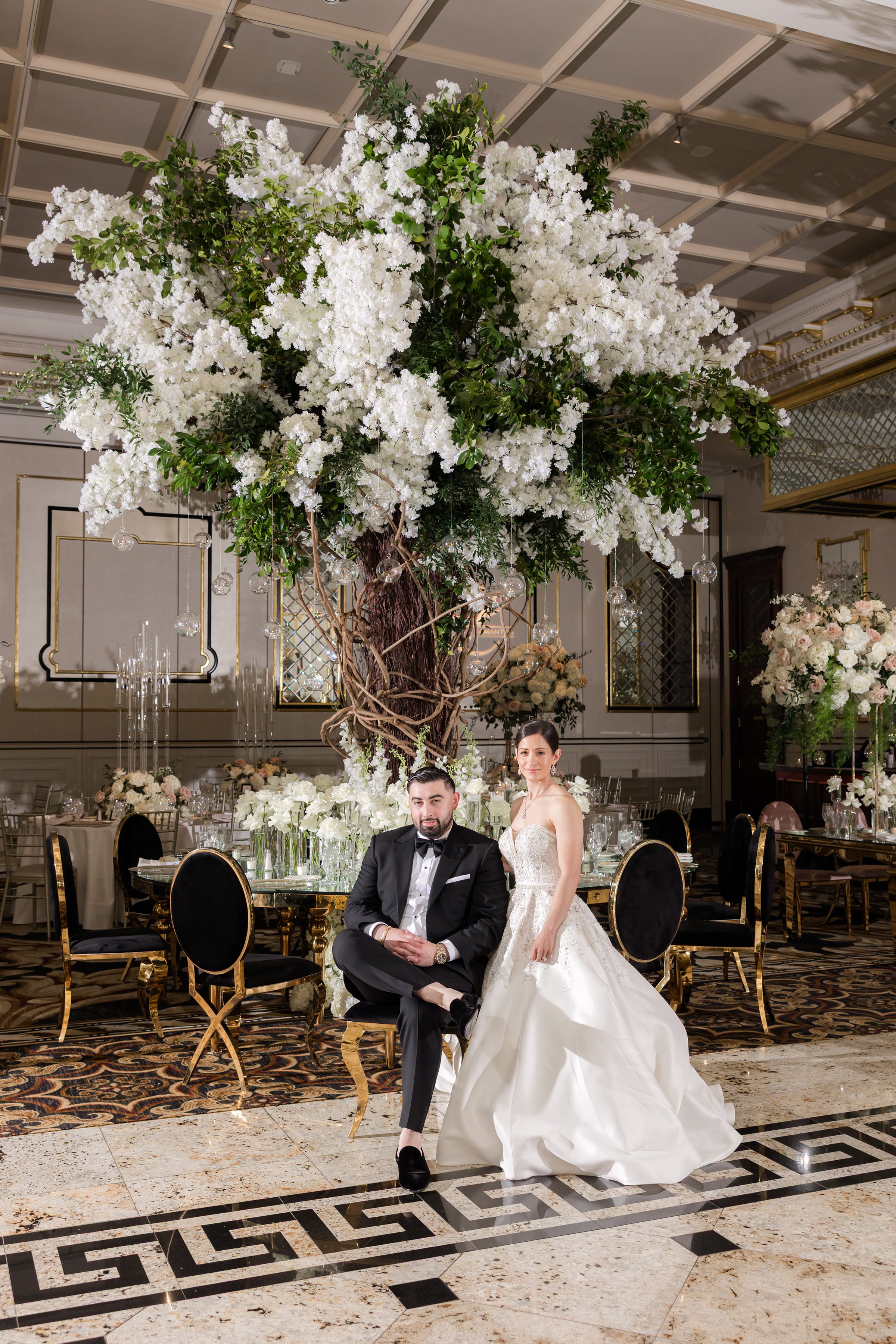 Portrait of groom sitting with legs crossed, and the bride standing, leaning into him. Behind them is the big tree centerpiece