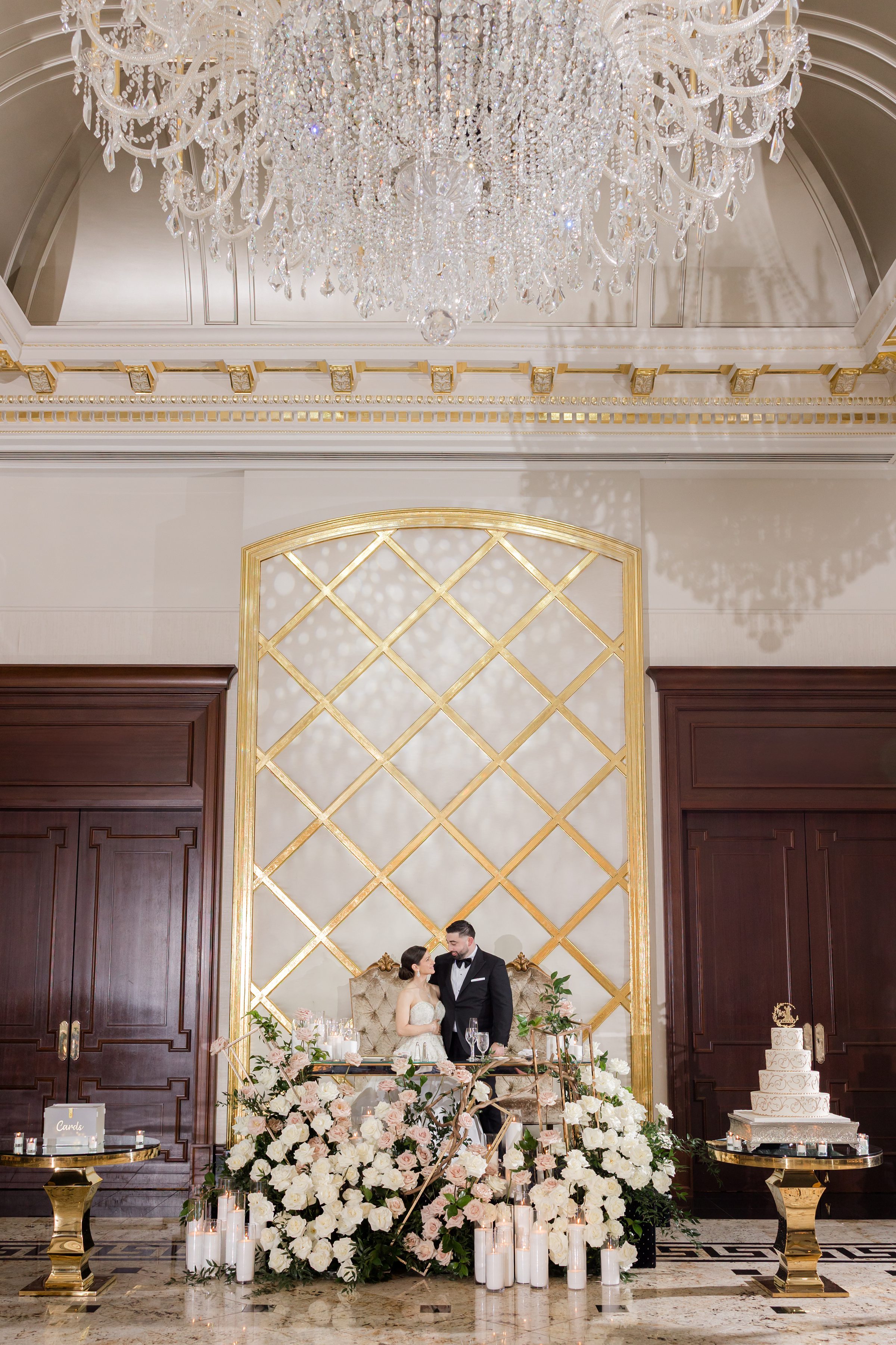 Bride and groom looking at each other at the sweetheard table, decorated with peach and white flowers, and candles. The the right side is their 4-tier cake, and behind is a white wall with gold decoration