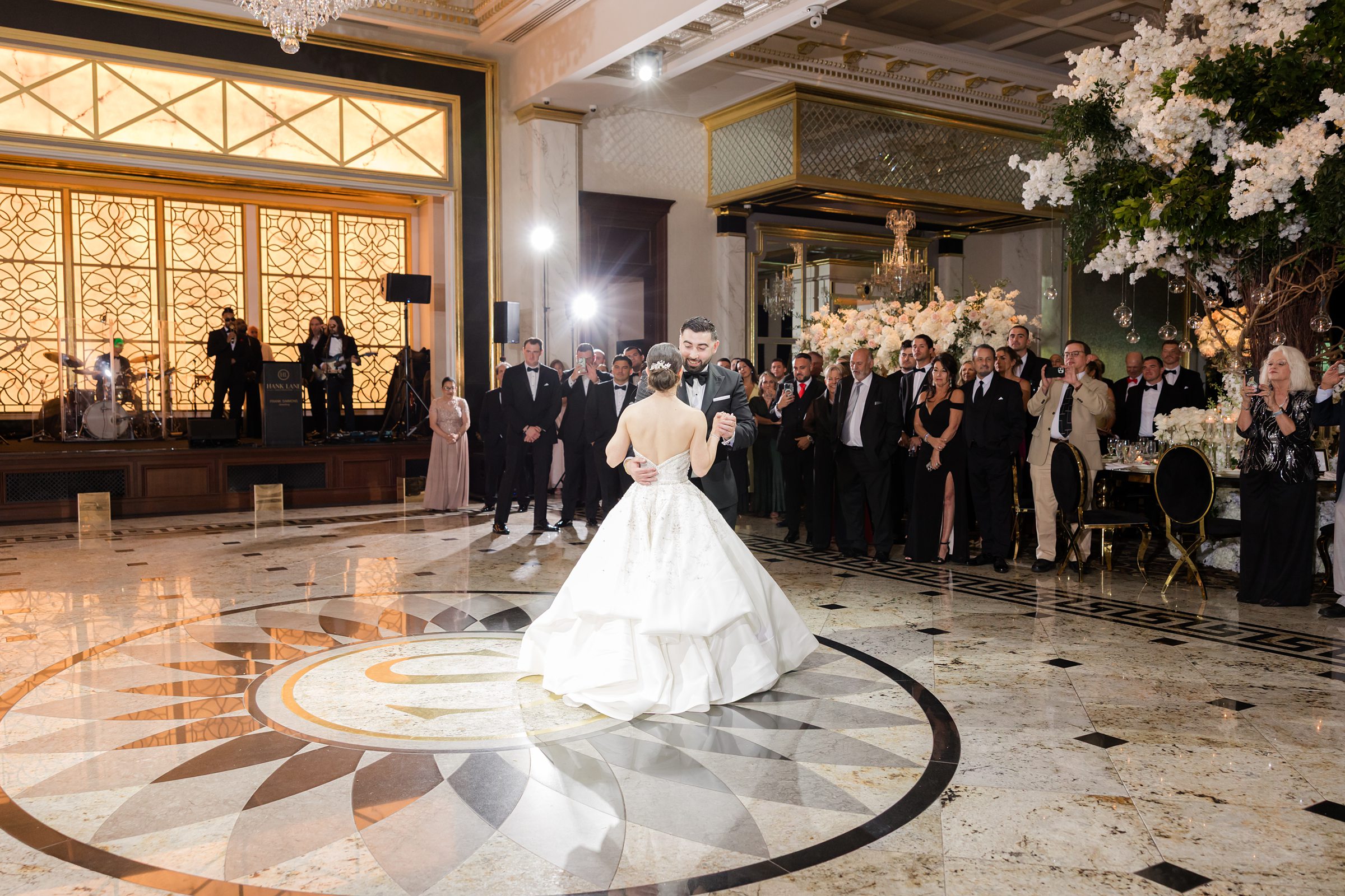Landscape photo of couple's first dance in a white, black, and gold ballroom, surrounded by their guests, with a live band