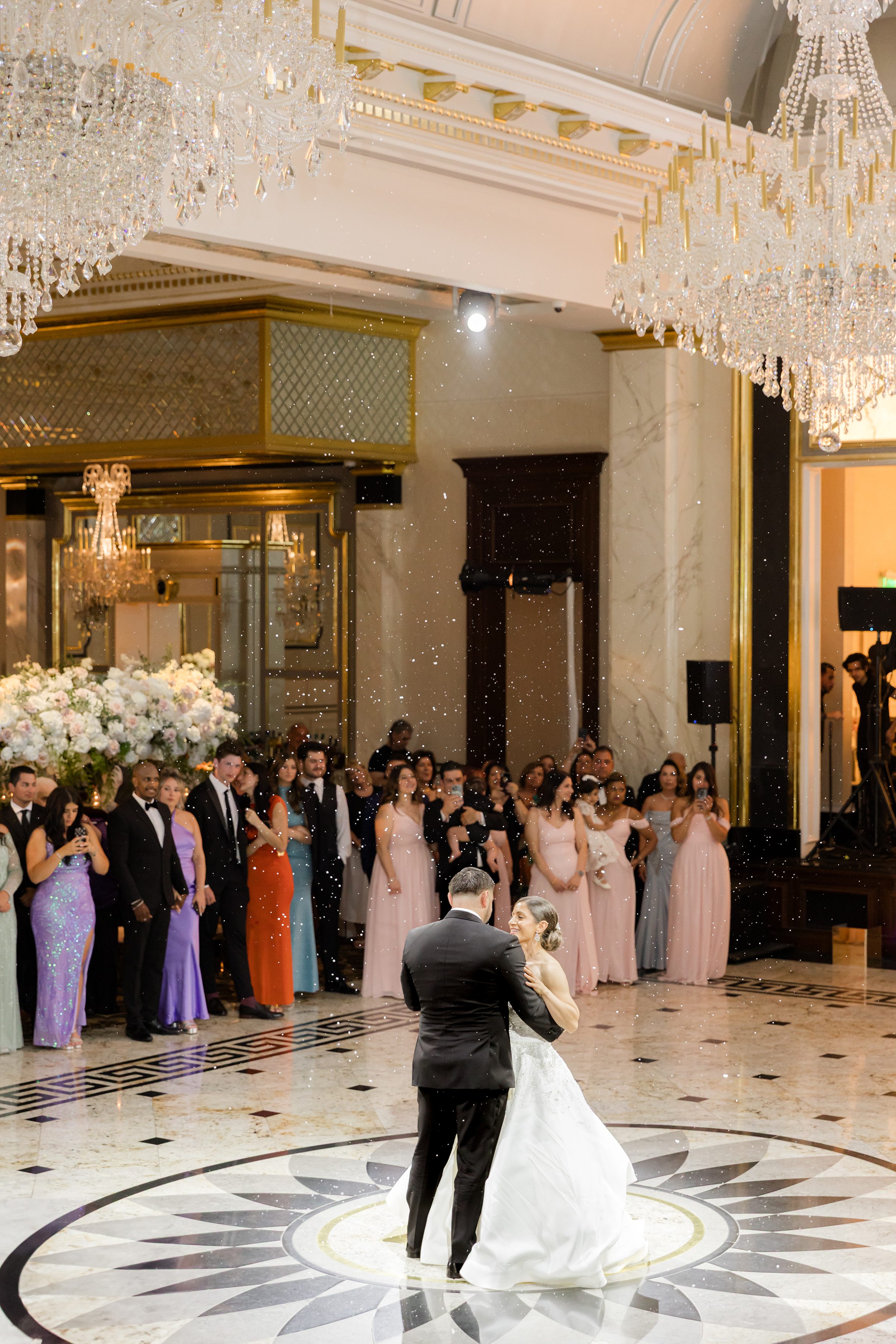 Portrait of bride and groom dancing while holding each other with white confetti falling down, while guests are watching them