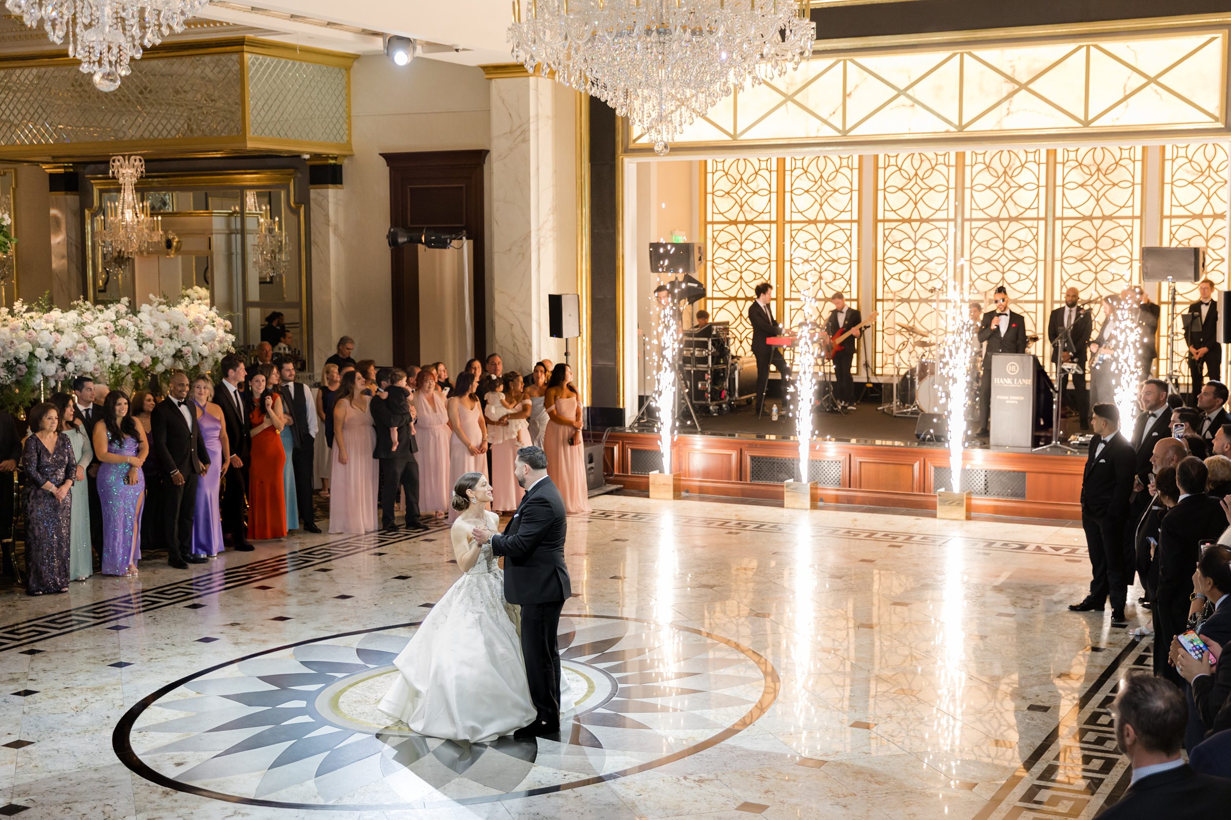 Landscape photo of bride and groom looking at each other as the guests surround them, with a live band playing and sparkler towers