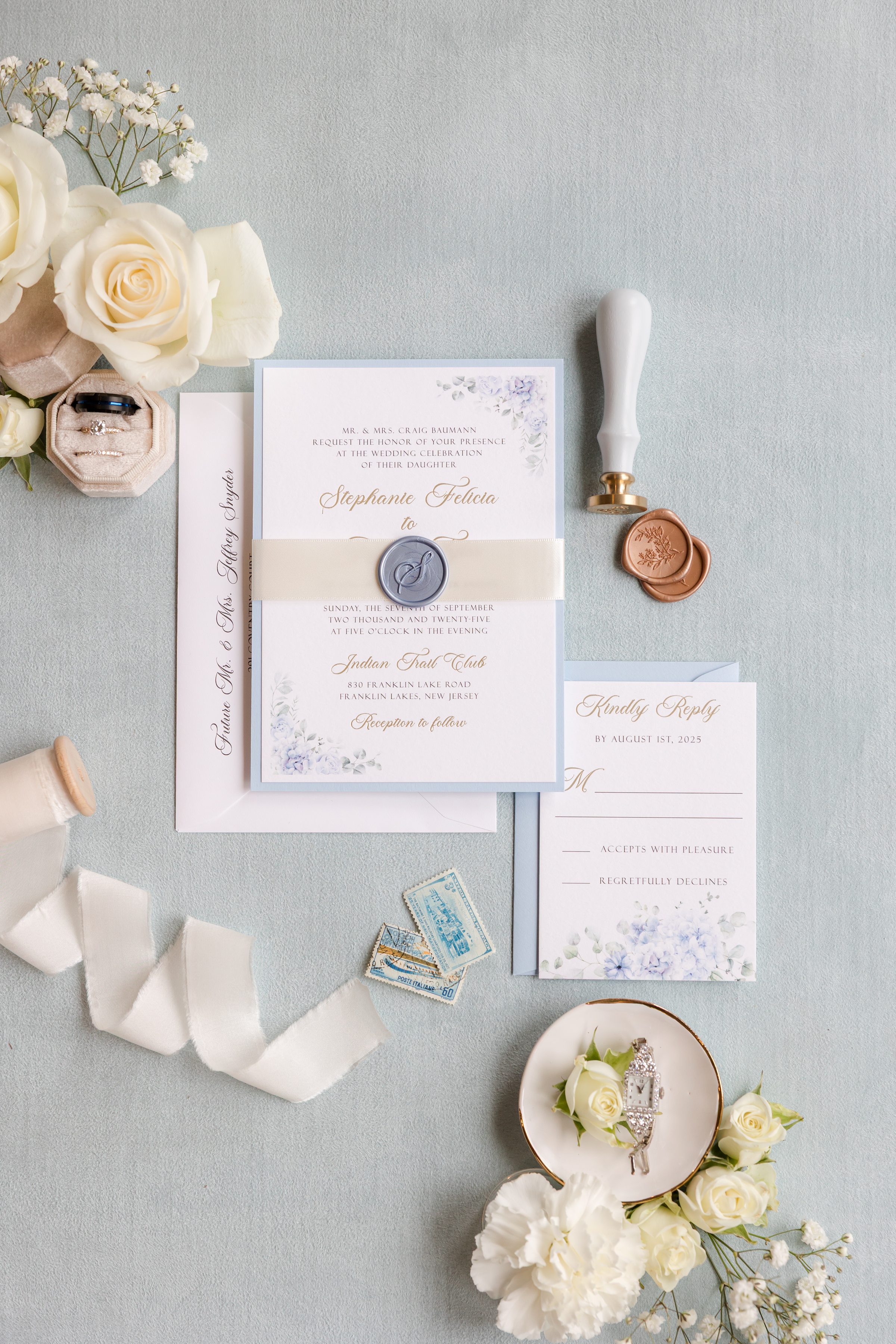 Elegant wedding stationery flat lay featuring an invitation suite tied with silk ribbon, a wax seal stamp, and soft ivory roses, beautifully arranged to capture timeless romance and refined bridal detail