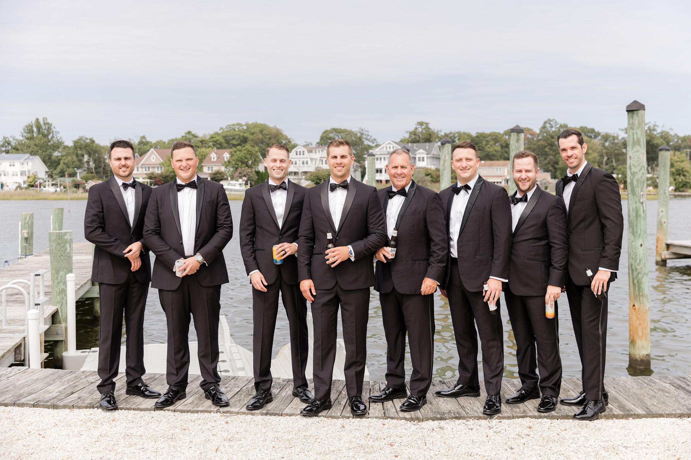 Groom stands with his groomsmen by the water, all in classic black tuxedos, sharing a relaxed and joyful moment before the celebration