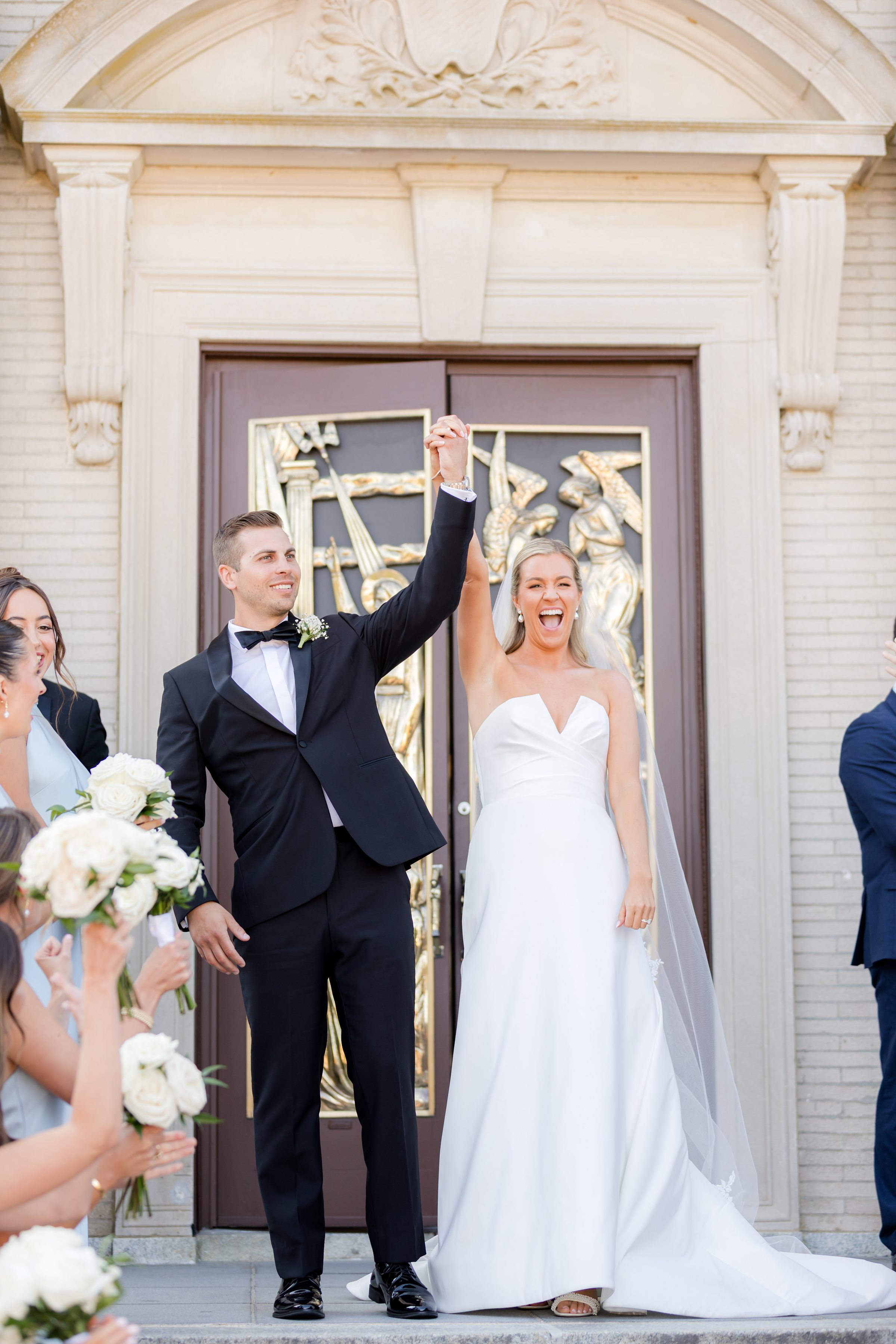 Newlyweds step out of the church together, raising their arms in celebration as joy and love surround them