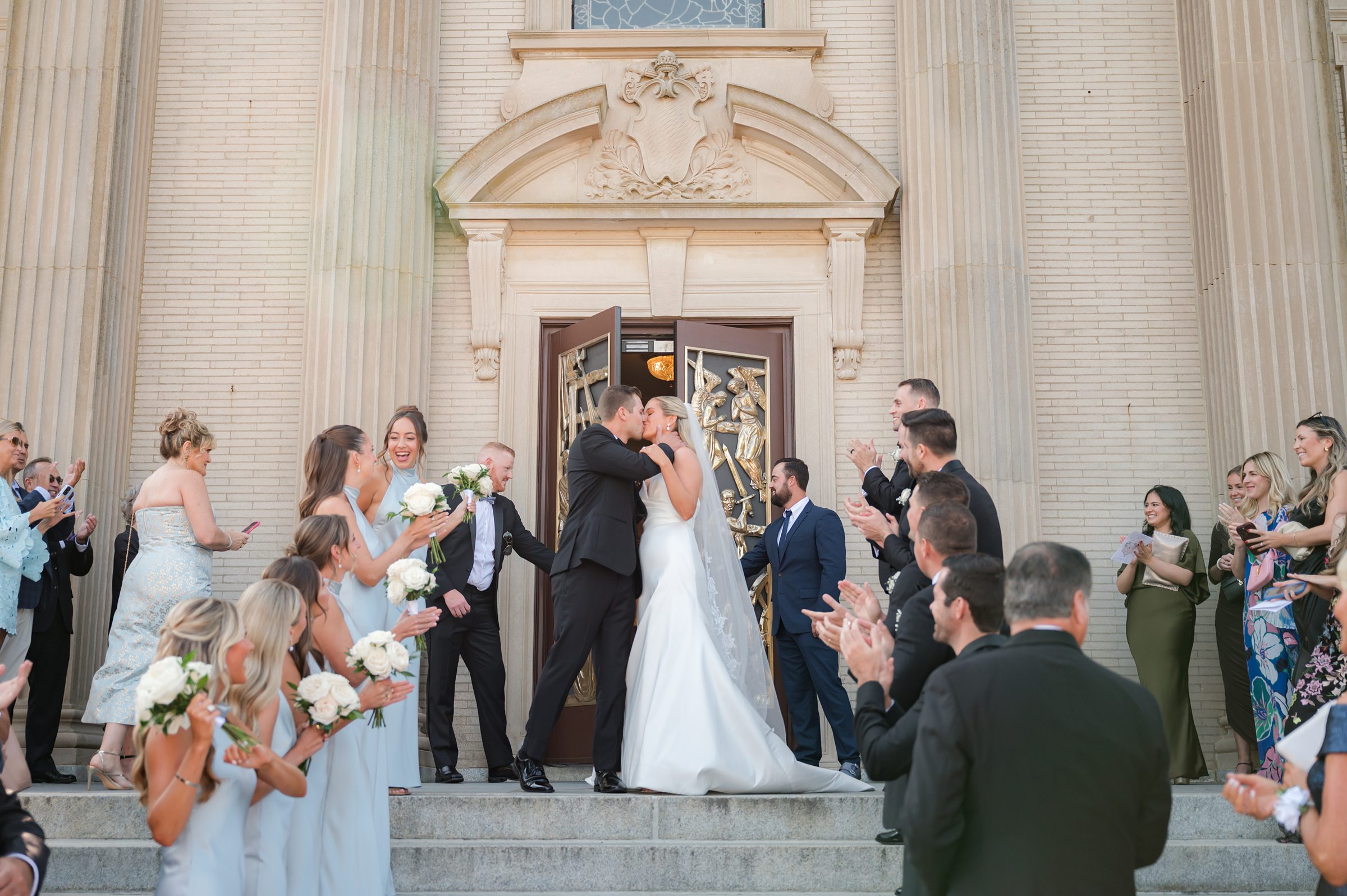 Newly married, the couple shares a joyful kiss on the church steps as loved ones cheer around them, marking the start of their life together.