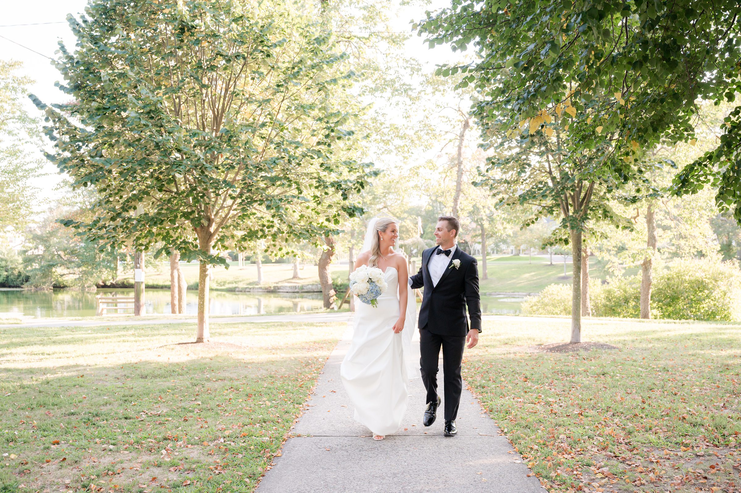 Bride and groom walk together along a tree-lined path, smiling at each other in a peaceful, sunlit park on their wedding day.