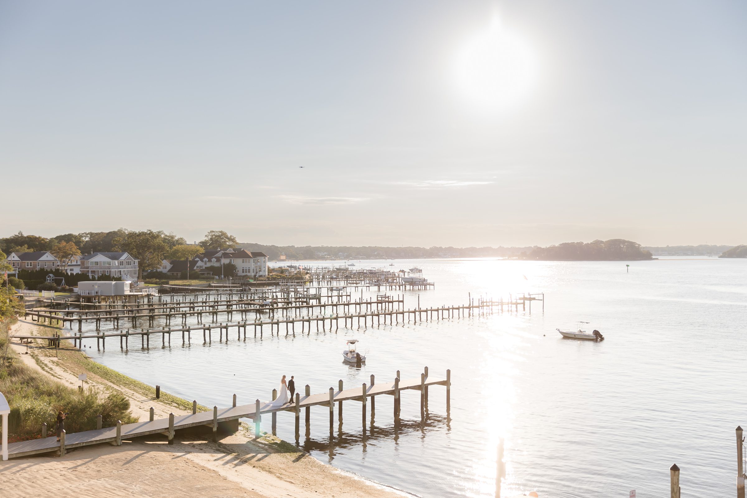 Sunset glows over a quiet harbor as a bride and groom walk together along a wooden dock, surrounded by calm water and soft light