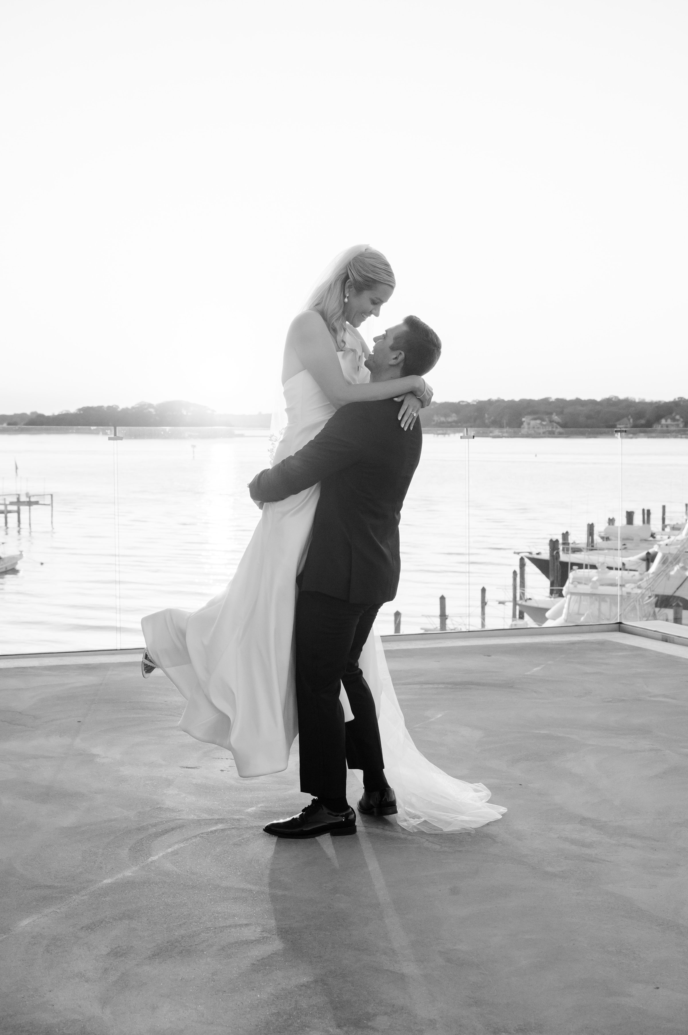 Groom lifts the bride into a loving embrace by the waterfront as they smile into each other’s eyes.
