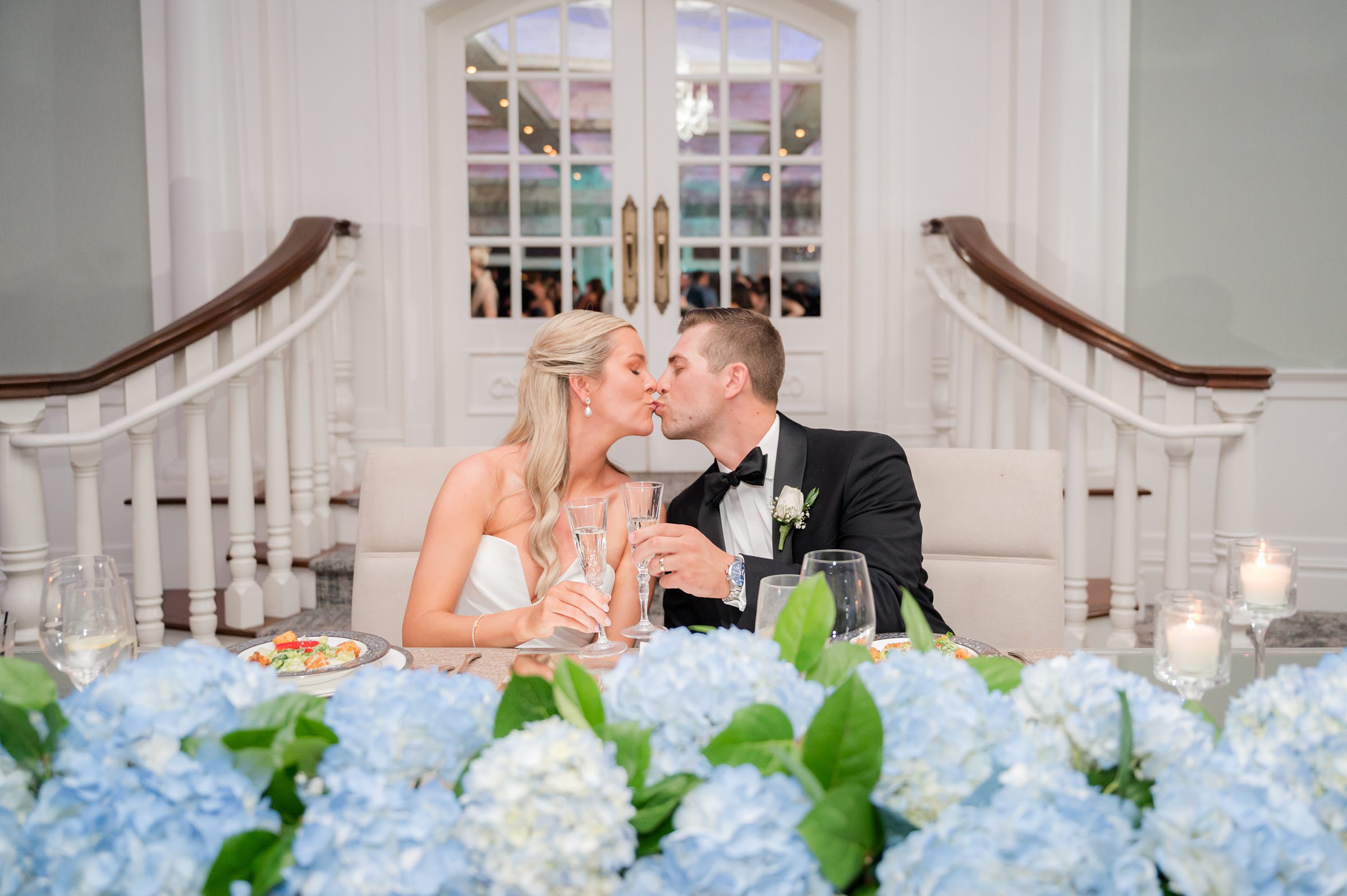 Bride and groom share a tender kiss over champagne, surrounded by soft candlelight and delicate blue florals.