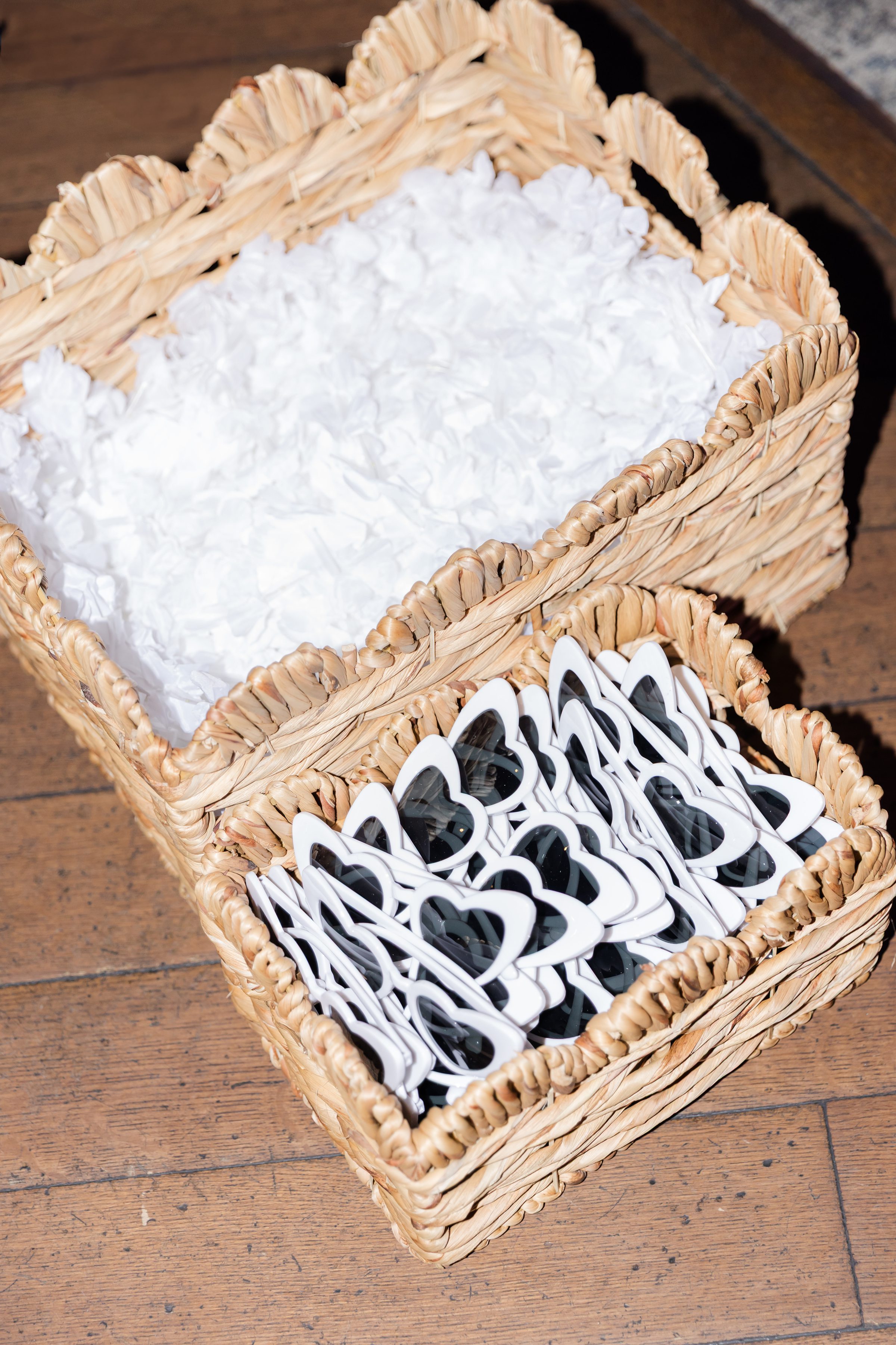 Woven baskets filled with white flower petals and heart-shaped sunglasses arranged for wedding guests.