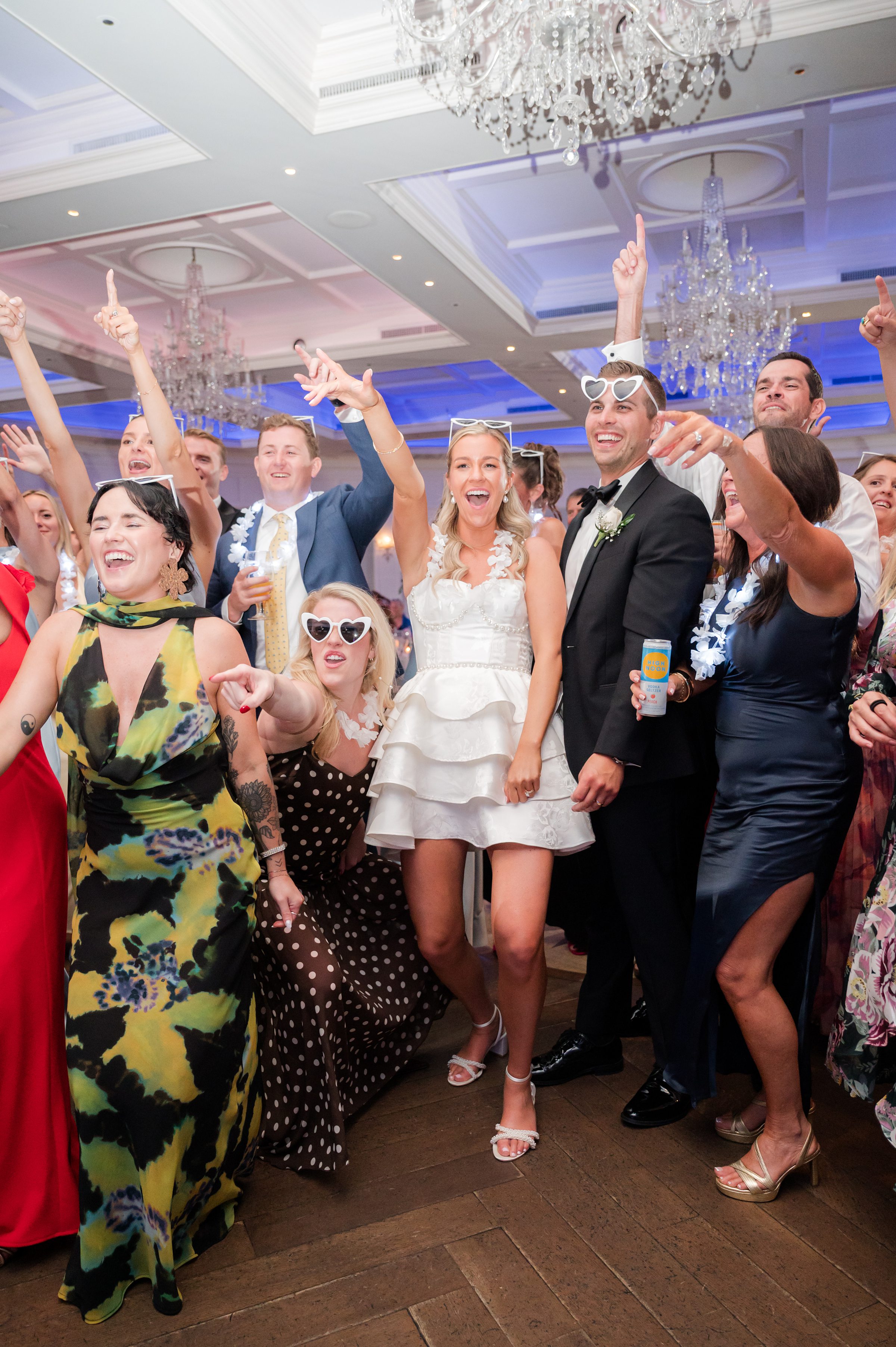 Bride and groom stand at the center of a lively crowd, smiling and celebrating as guests dance and cheer around them