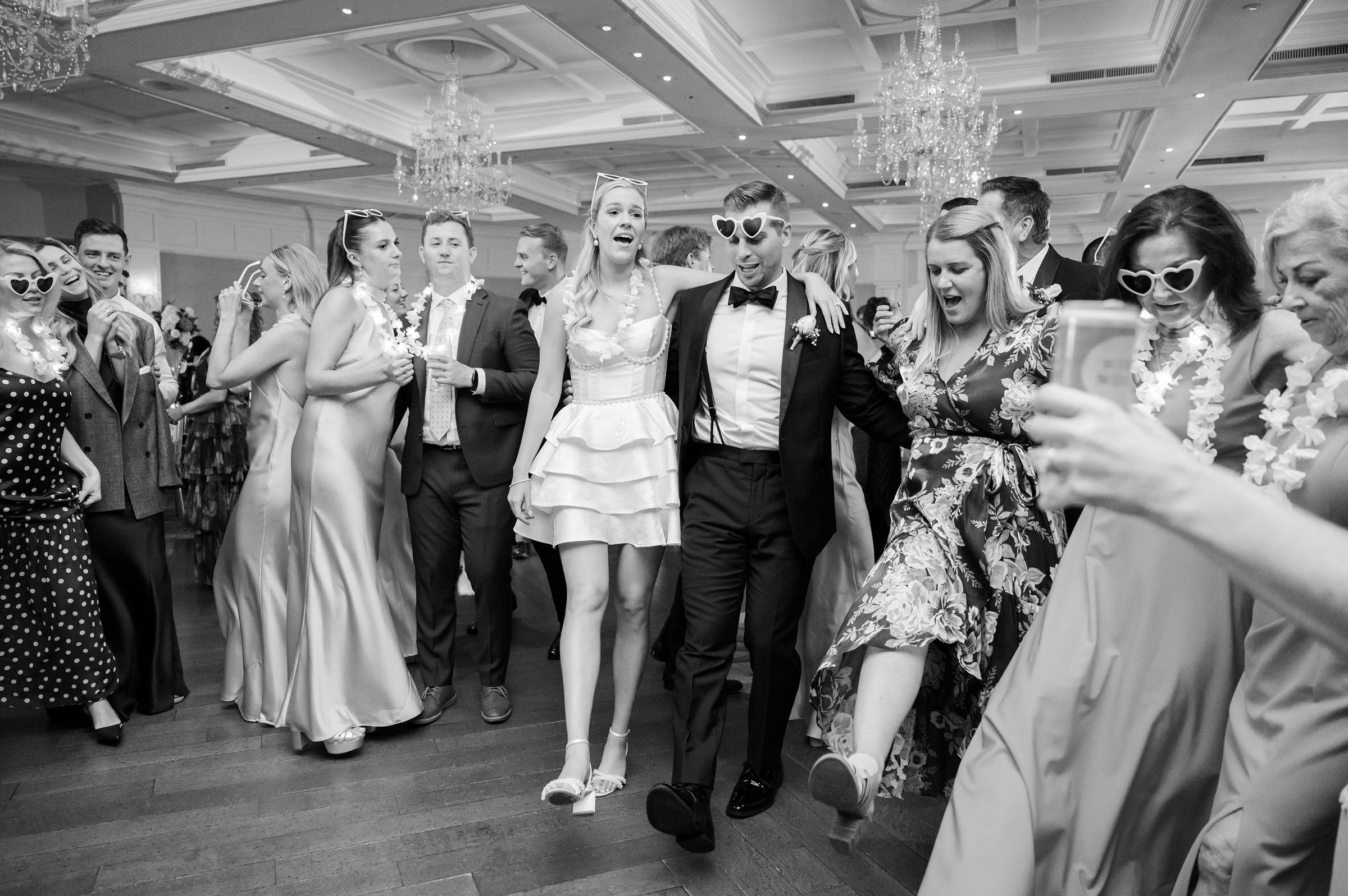 Bride and groom share a carefree moment as they walk together, surrounded by dancing friends and laughter.
