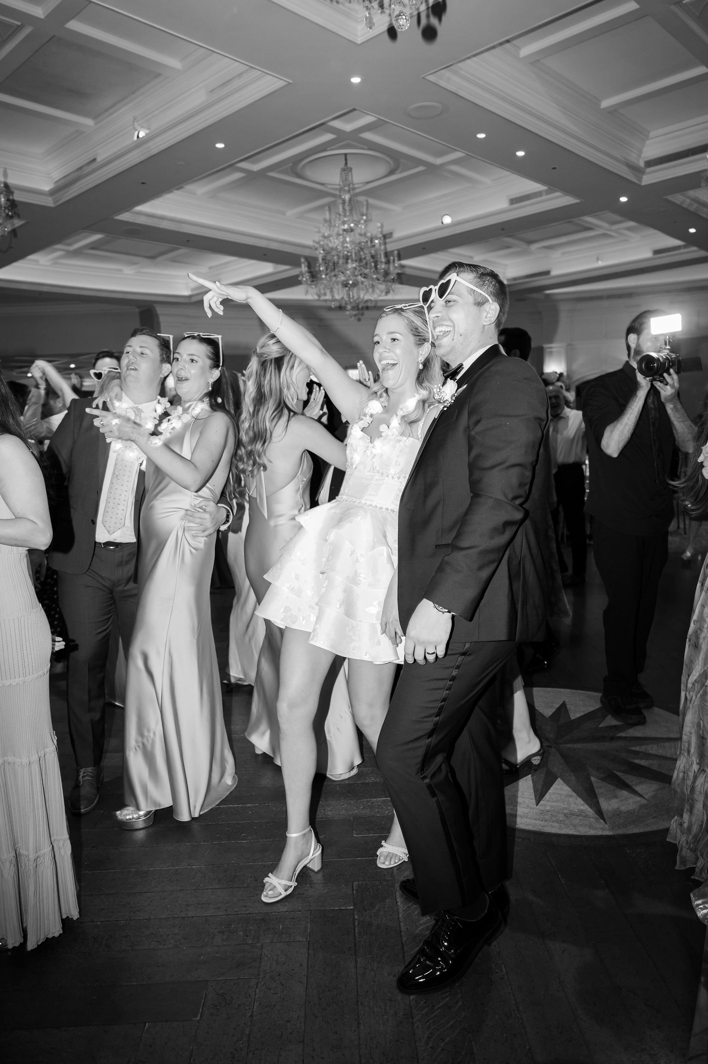 Bride points joyfully across the dance floor while the groom stands beside her, both laughing and fully immersed in the celebration.