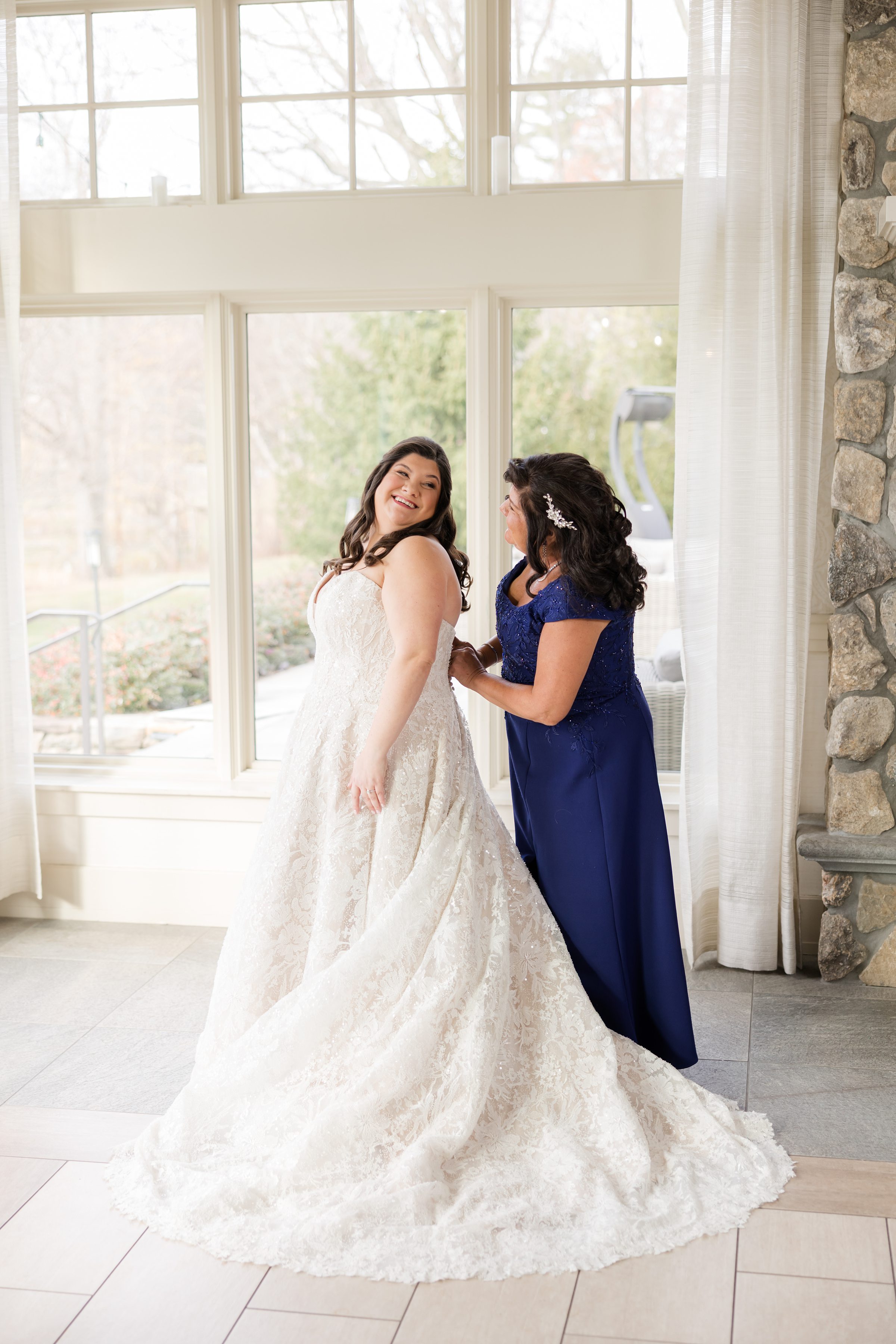 Bride smiling in a lace wedding gown as her mother helps fasten the back of the dress in a sunlit room
