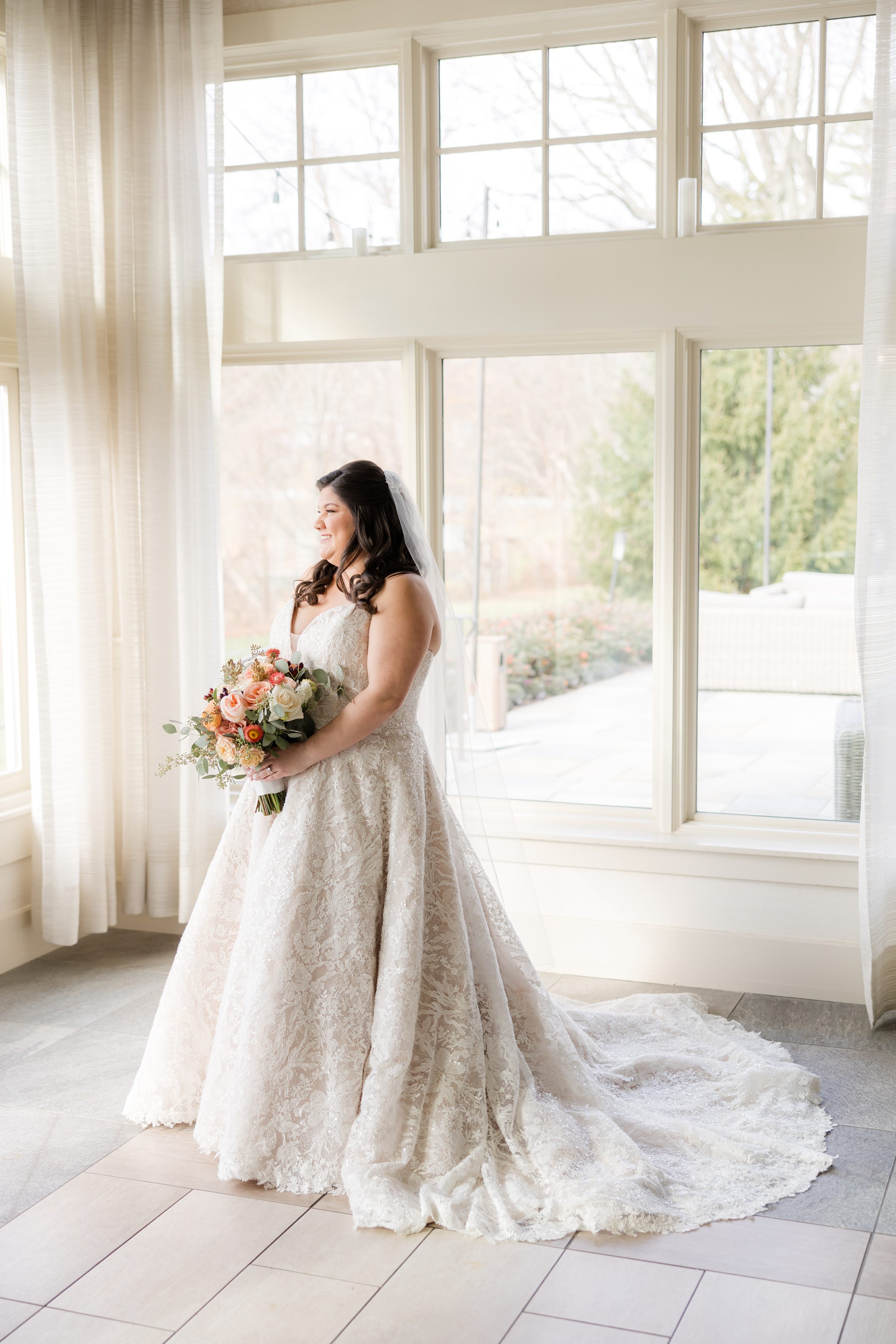 Bride in a lace wedding gown holding a bouquet of peach and white flowers, standing by large sunlit windows