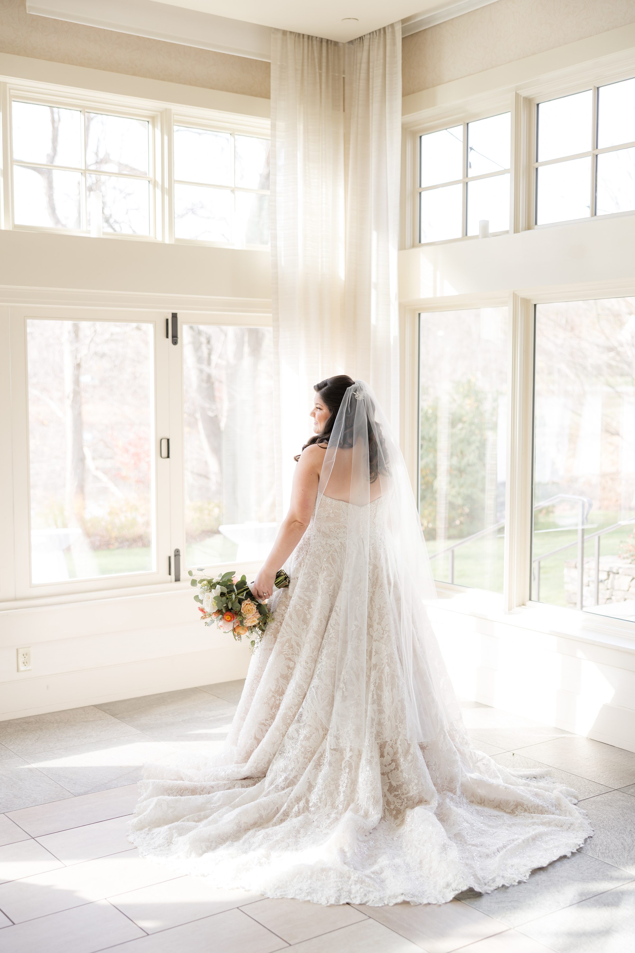 Bride in a lace gown holding a bouquet, standing in a bright room with tall windows and soft natural light
