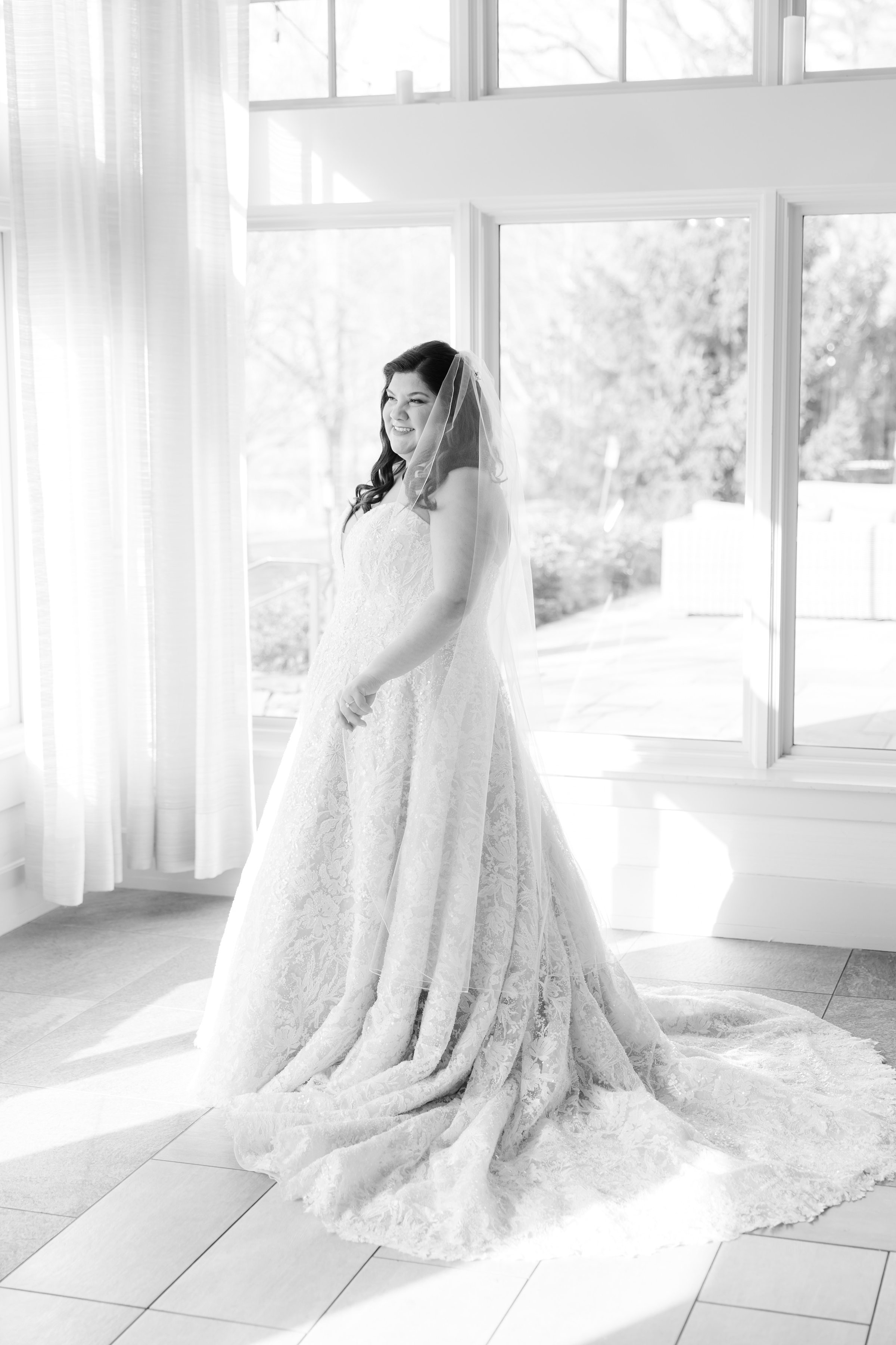 Bride in a lace wedding gown and veil standing by large sunlit windows in a softly lit room, smiling