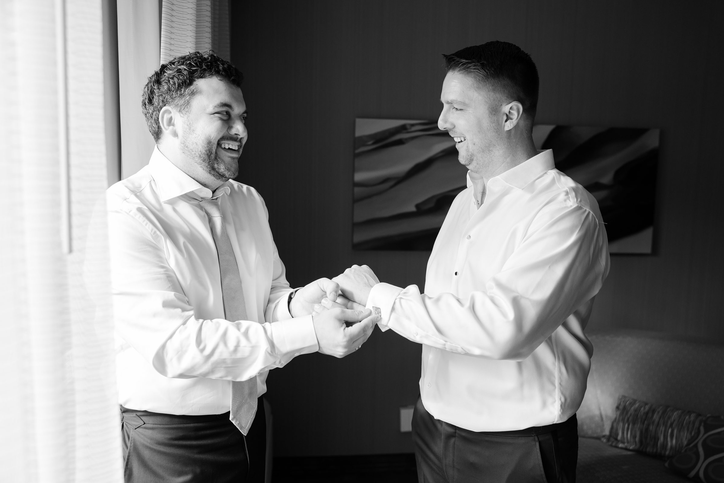 Groom and groomsman laughing while fastening cufflinks in a black and white candid shot