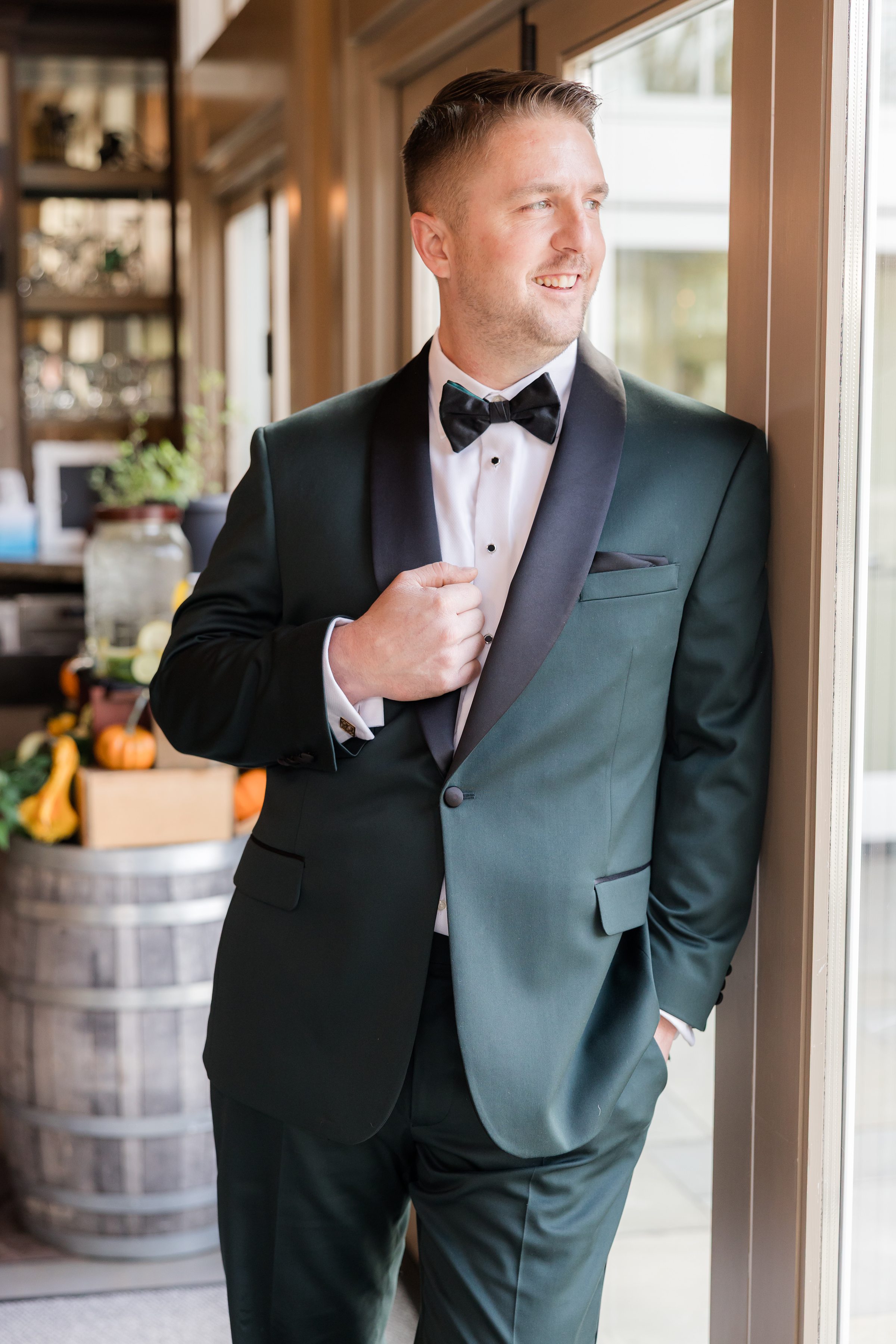 Groom in green tuxedo standing by a window indoors, looking out thoughtfully