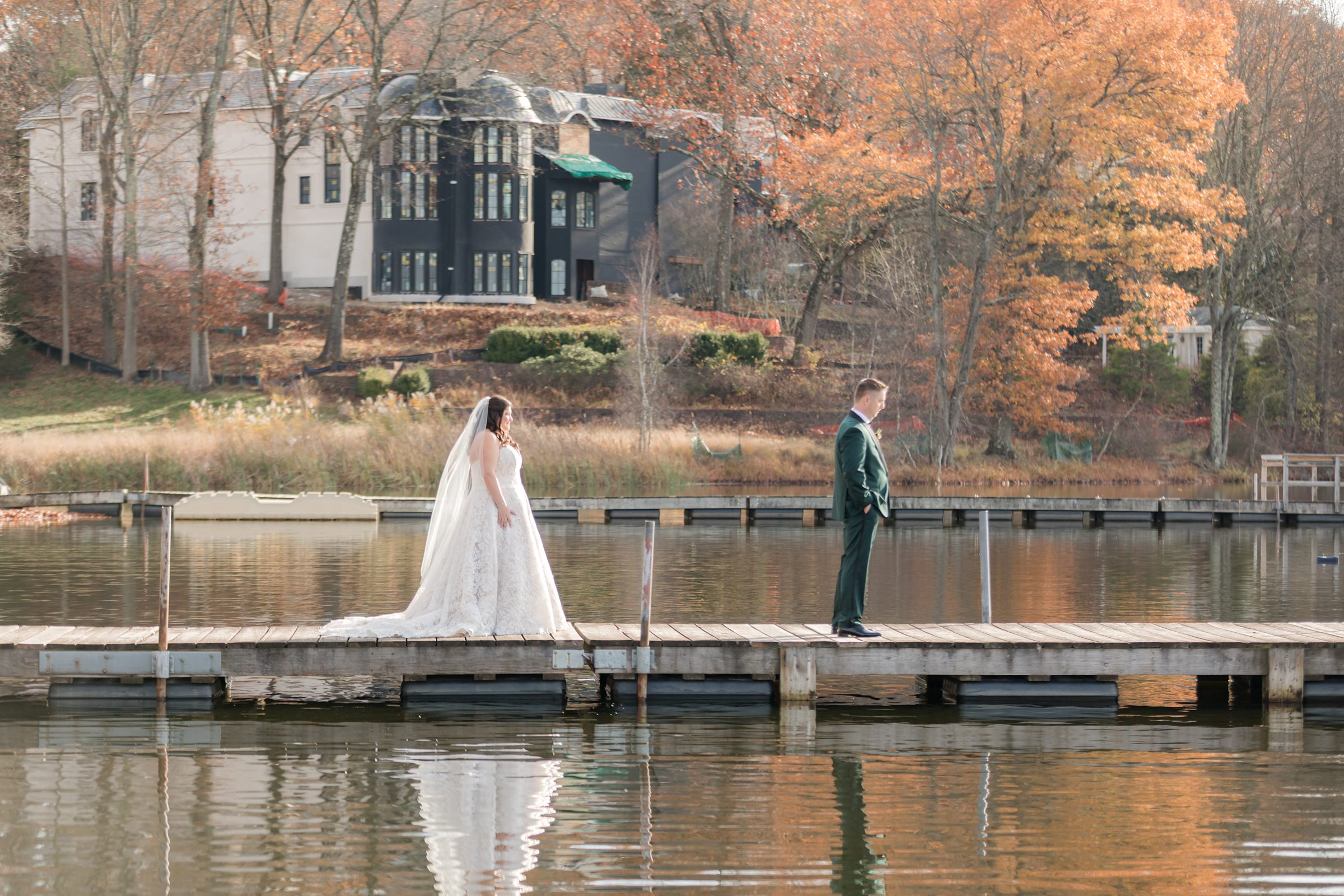 Bride and groom standing apart on a dock over calm water with fall trees in background