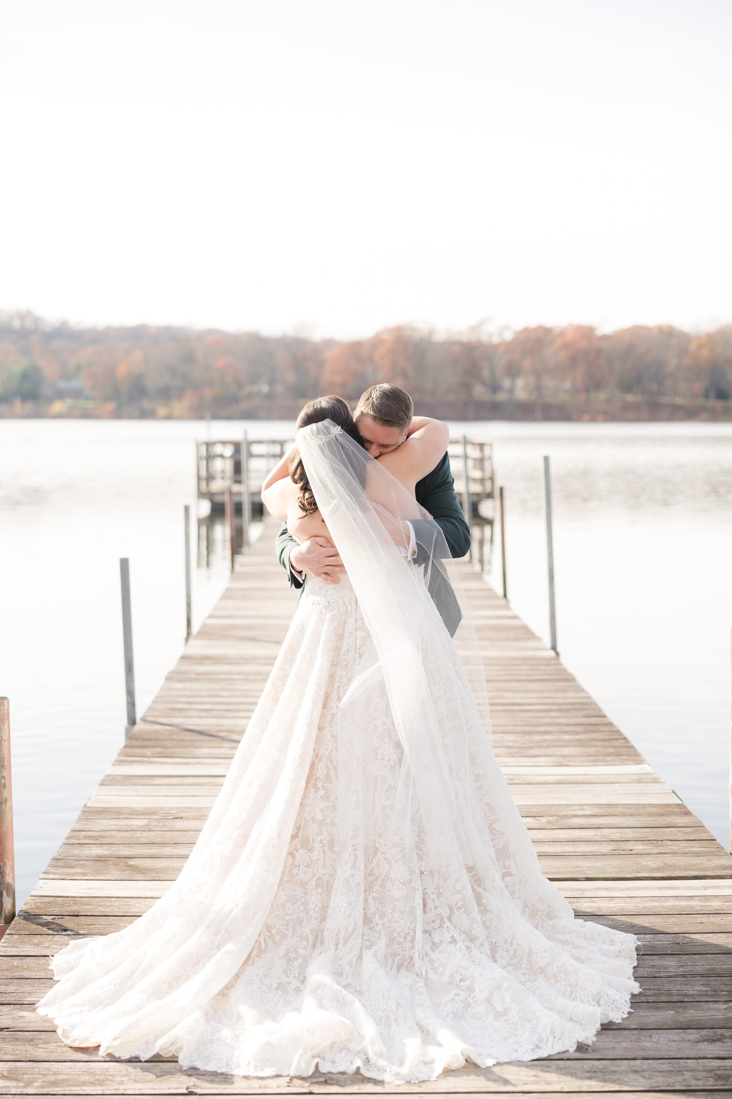 Bride and groom embracing tightly on a dock bride’s long veil flowing behind.