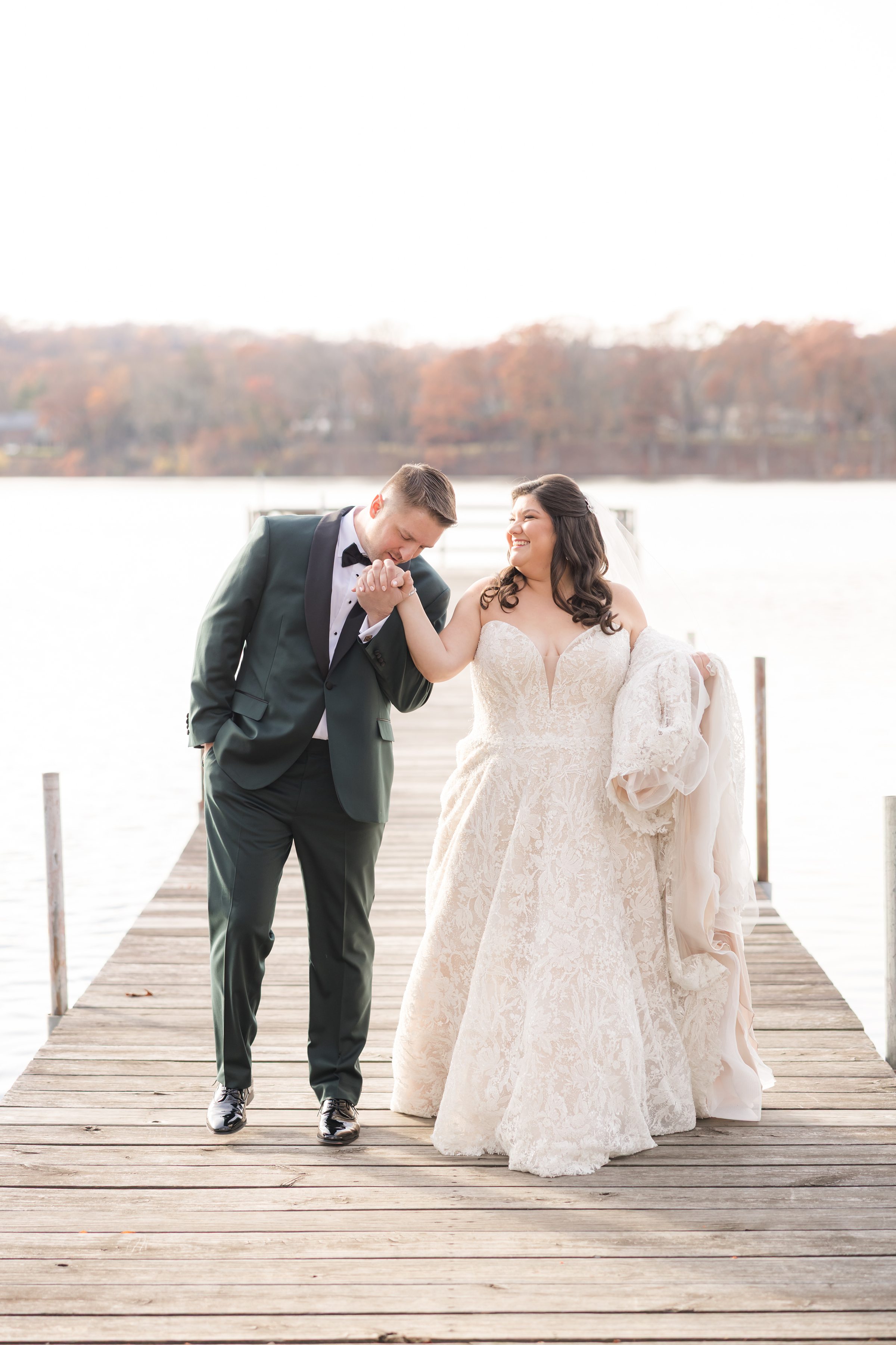 Groom kisses bride’s hand as they walk together on a lakeside dock