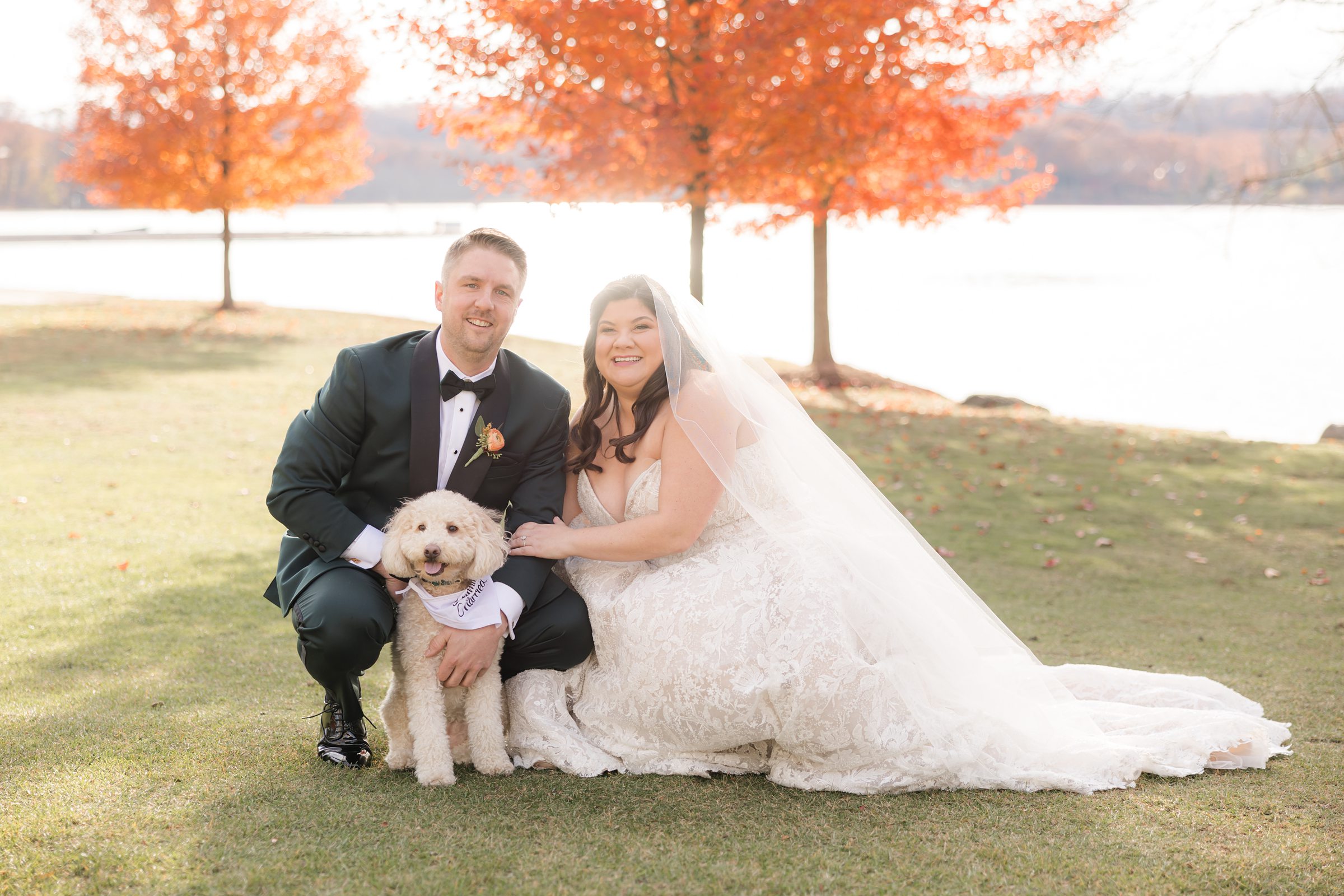 Bride and groom kneeling with their dog between them, smiling near a lakeside with orange trees