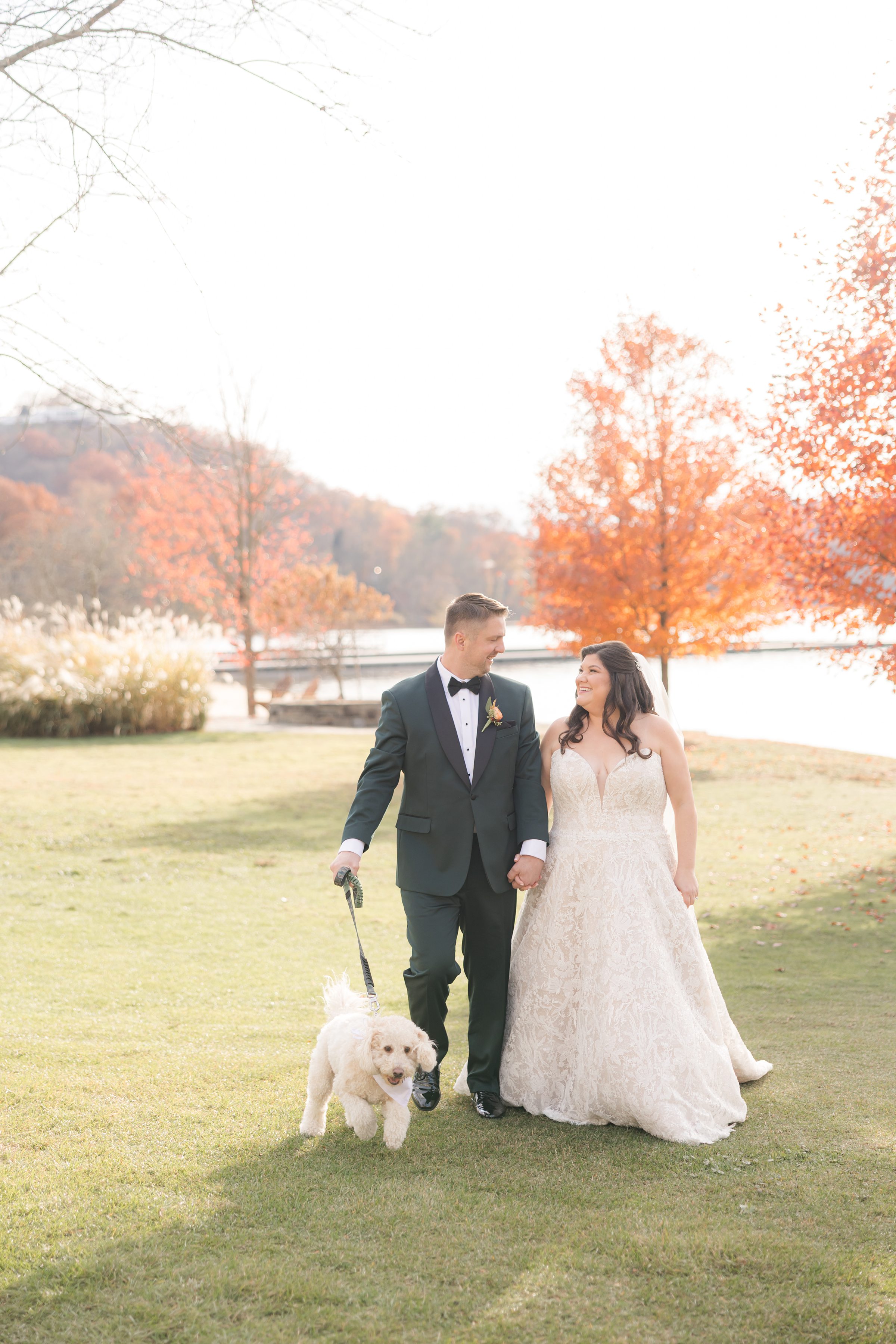 Bride and groom walk together along the lakeside, smiling at each other beneath glowing autumn trees