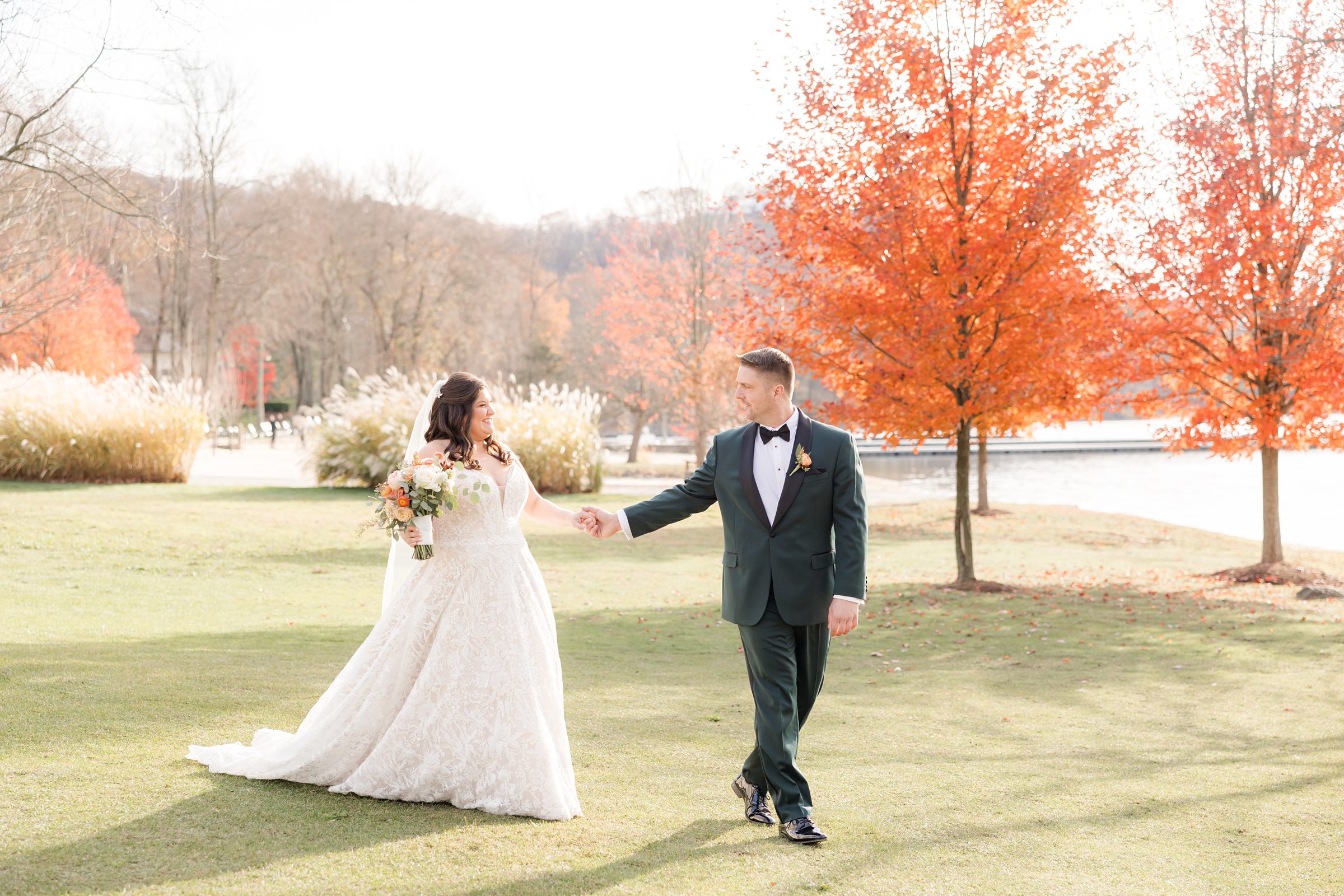 Bride and groom holding hands and walking across a lawn with bright autumn trees by a lake