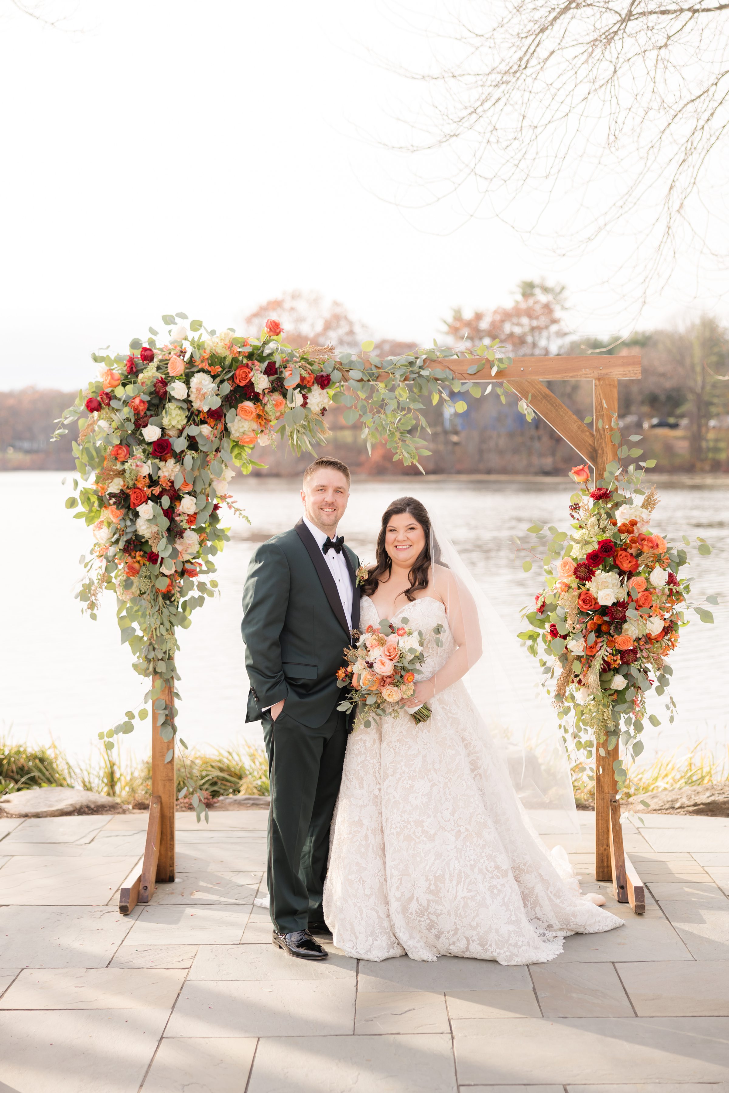 Bride and groom standing together under a wooden arch decorated with colorful flowers by the water