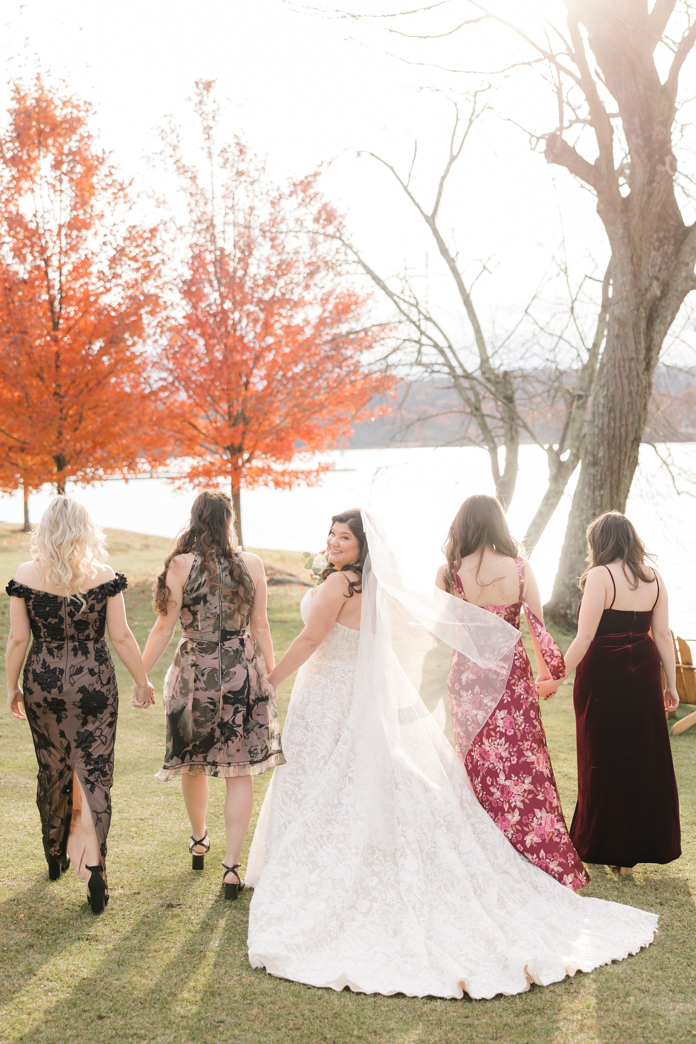 Bride and bridesmaids walking away together on grass near a lake, fall foliage in background