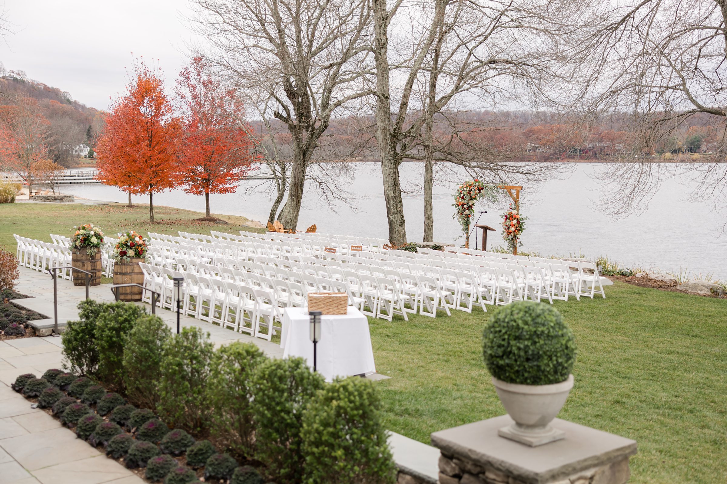 Outdoor wedding ceremony setup with rows of white chairs on a lawn overlooking a lake, featuring a floral arch at the altar and vibrant autumn trees in the background