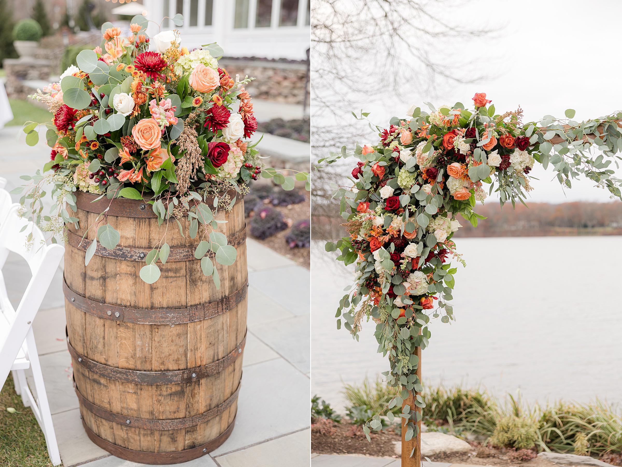 Rustic wooden barrel topped with a lush arrangement of roses, greenery, and fall-toned flowers alongside a floral ceremony arch decorated with cascading greenery and orange, red, and cream blooms by the lakeside