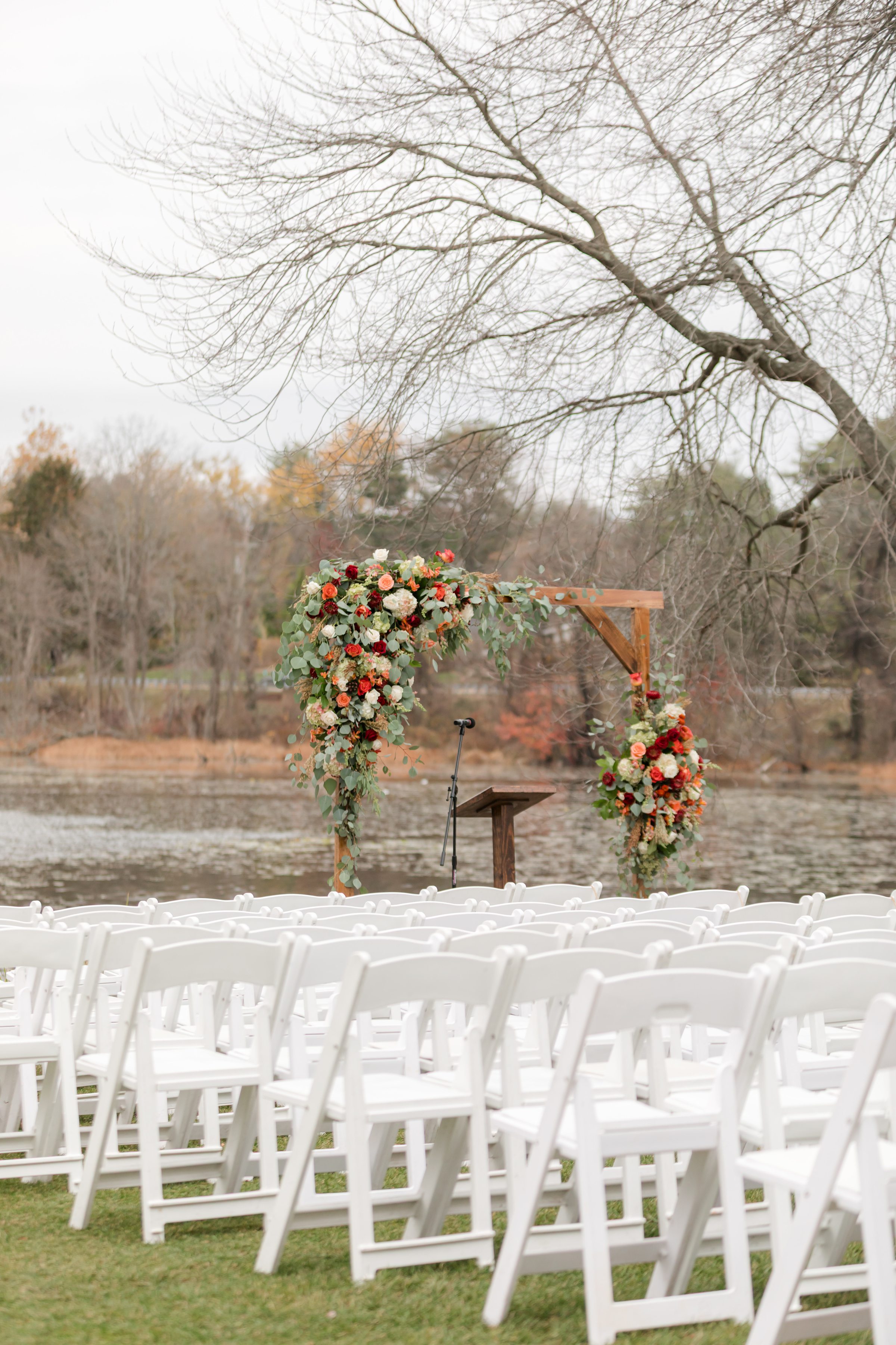 Outdoor wedding ceremony altar with a wooden arch adorned in cascading greenery and warm toned florals, set by a lakeside with rows of white chairs in the foreground and bare trees in the background
