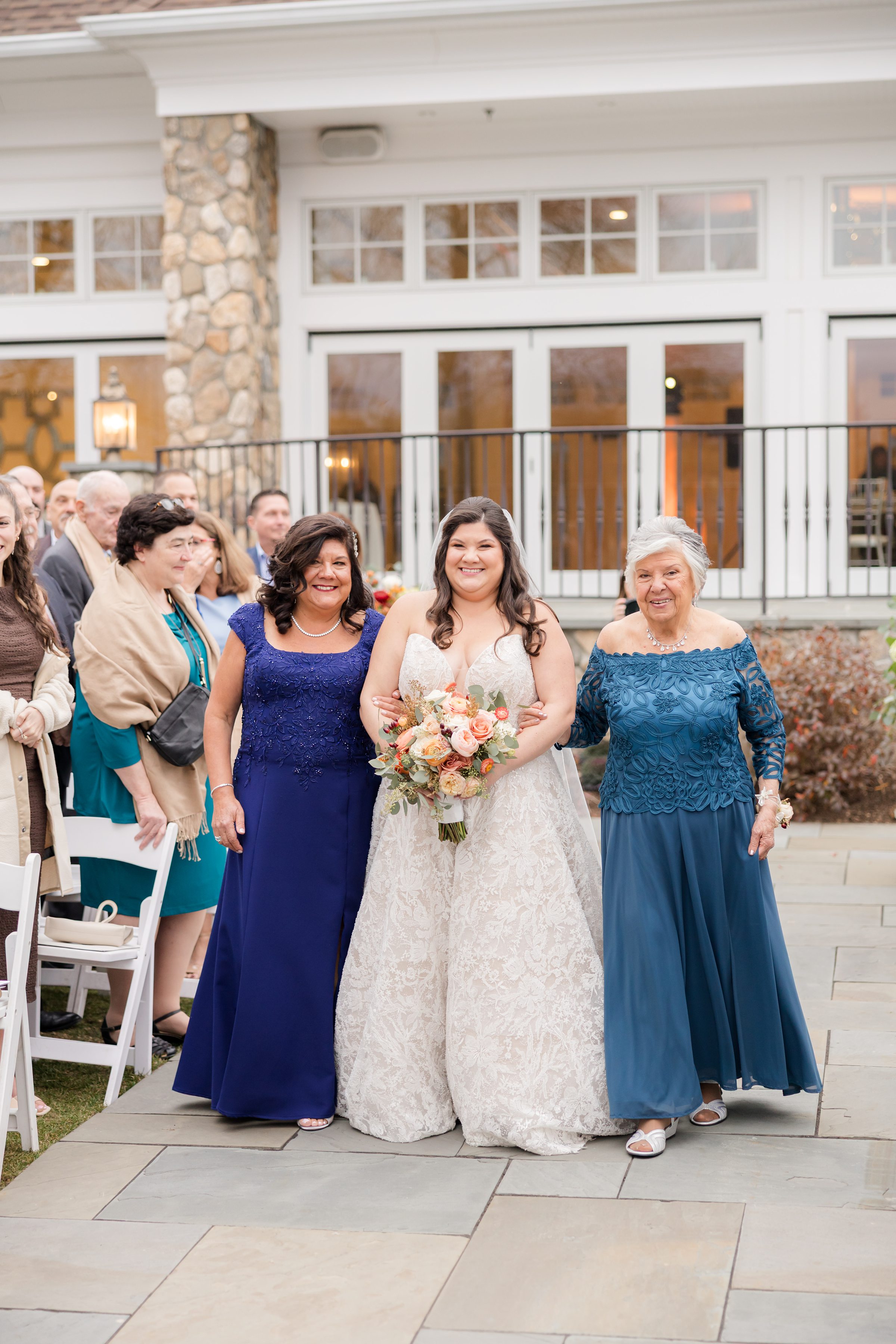 Bride walking down the aisle outdoors, smiling and holding a bouquet, escorted by her mother and grandmother, with guests watching in the background