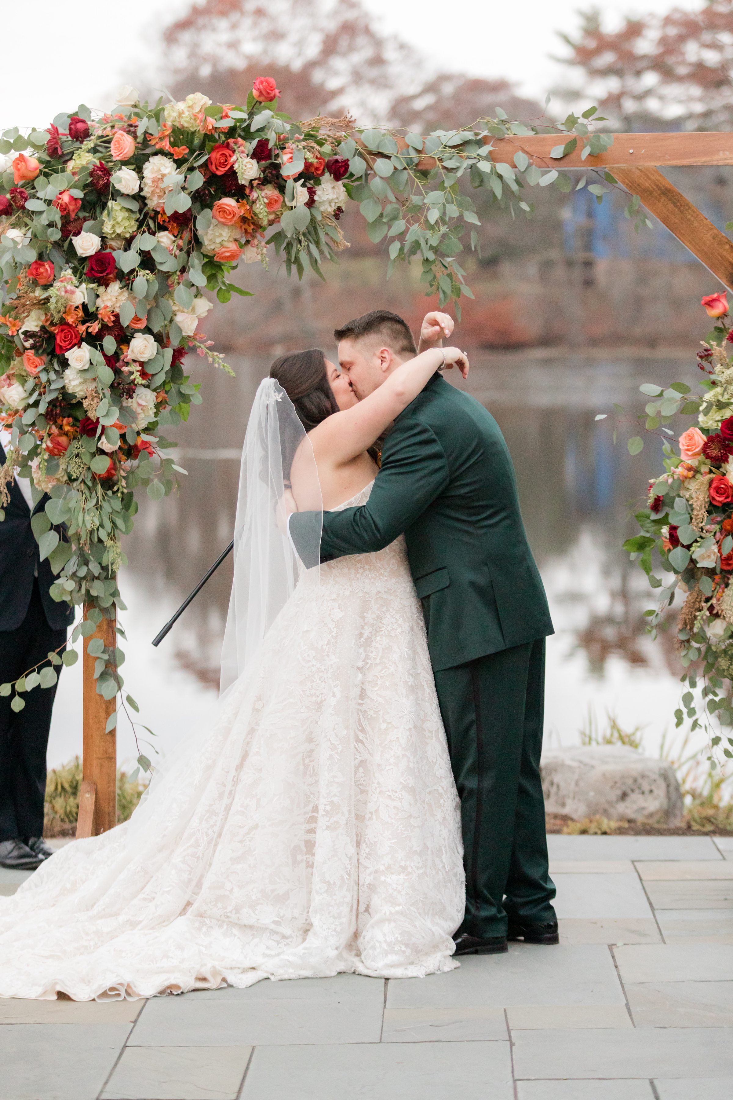 Bride and groom kissing under a floral arch by the water, surrounded by greenery and autumn tones