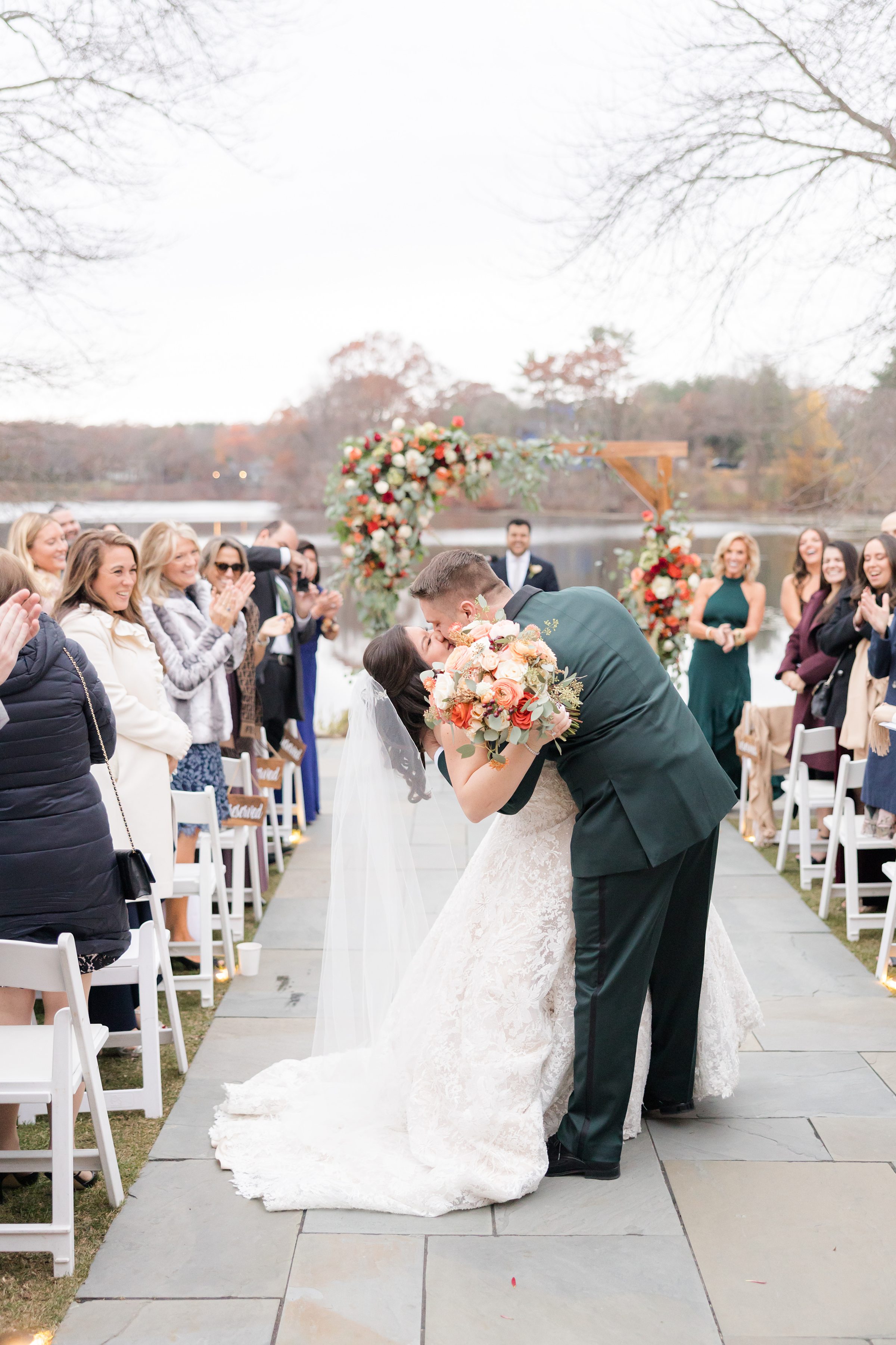 Groom dips the bride into a romantic kiss in the aisle as she holds a bouquet of soft peach and cream flowers, her lace gown and veil flowing, while guests on both sides smile and applaud