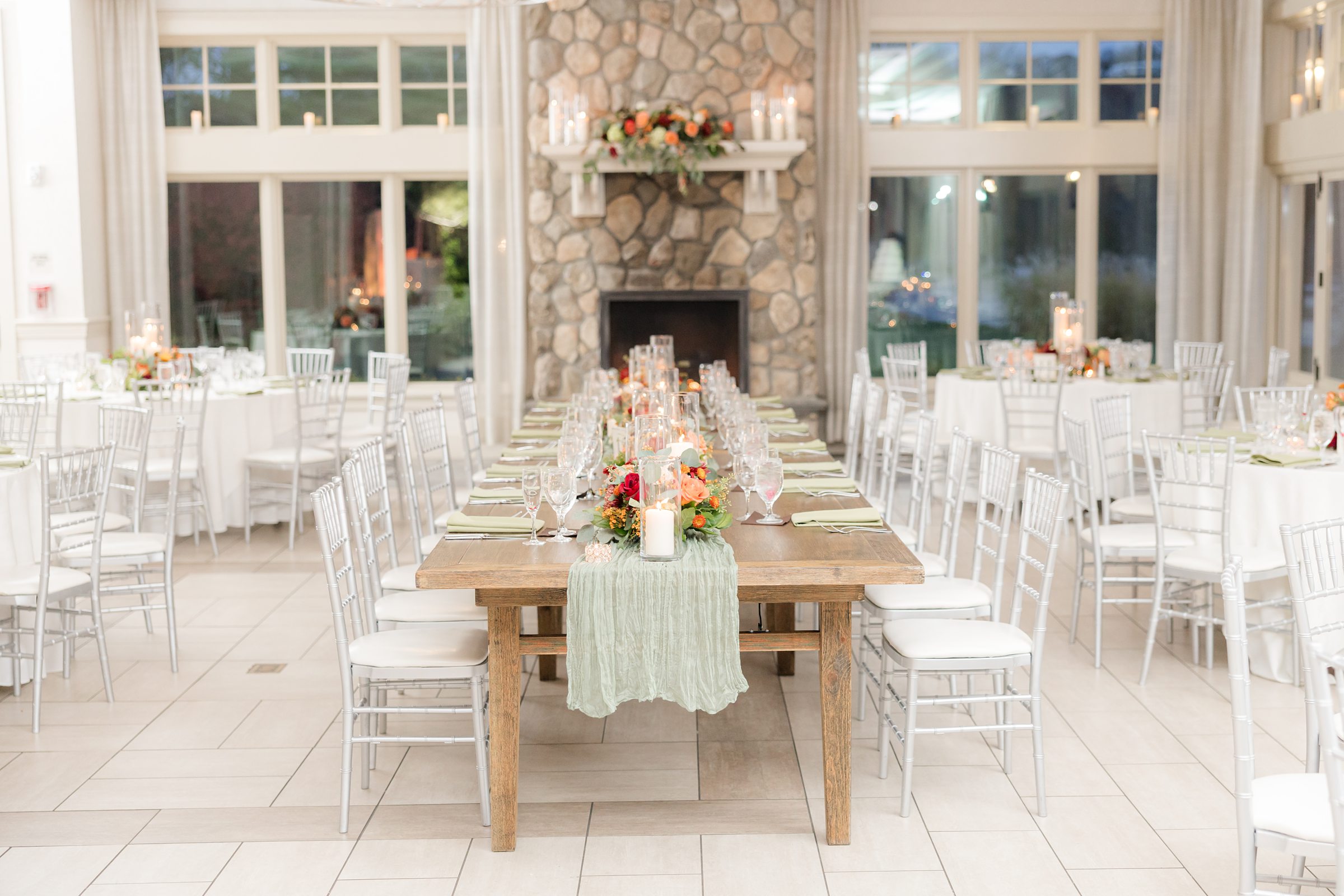 Long wooden reception table with candles and greenery runner, surrounded by white chairs in a bright, airy ballroom with a stone fireplace