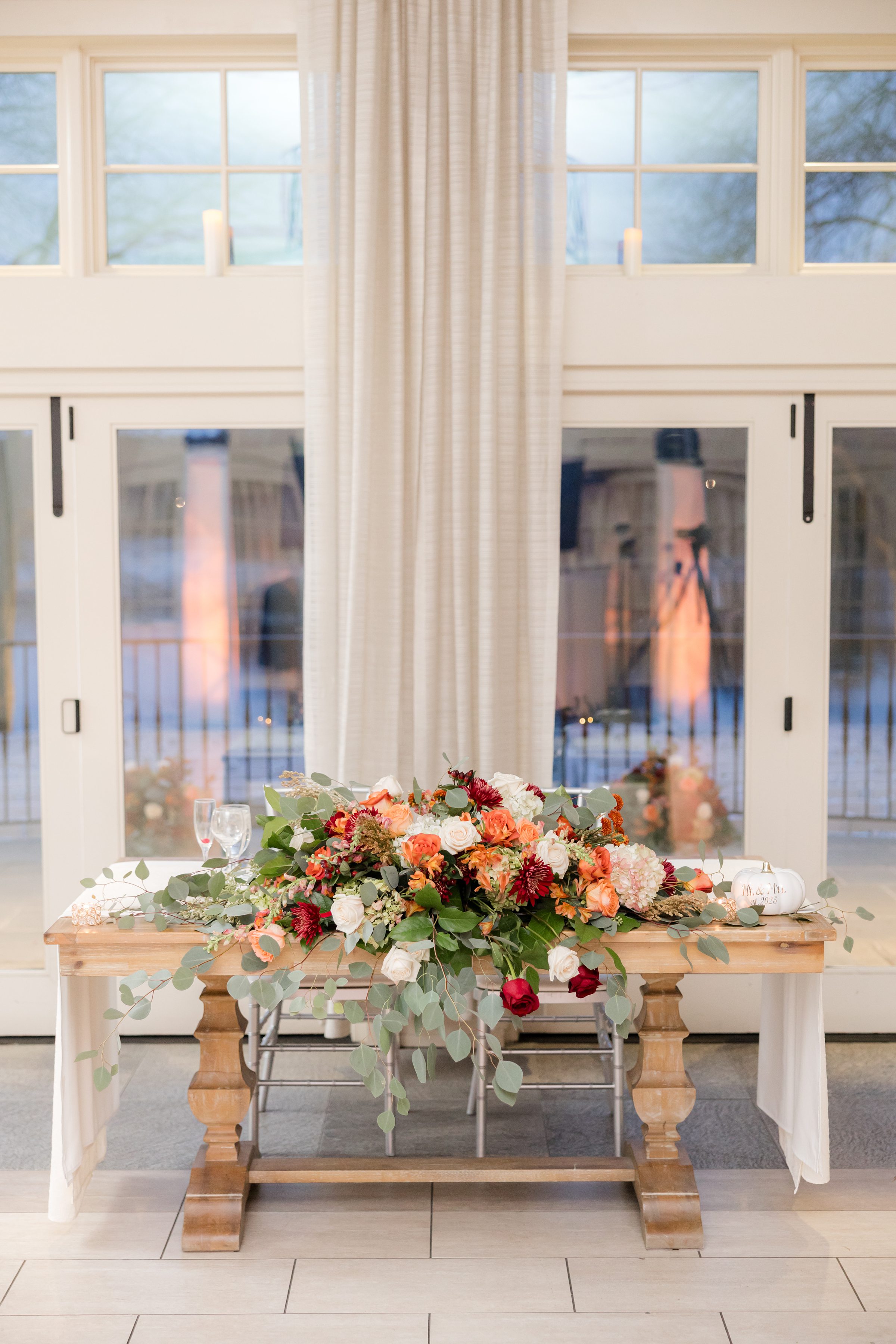 Elegant wedding sweetheart table with a lush floral arrangement in warm tones, set in front of tall windows and soft drapery