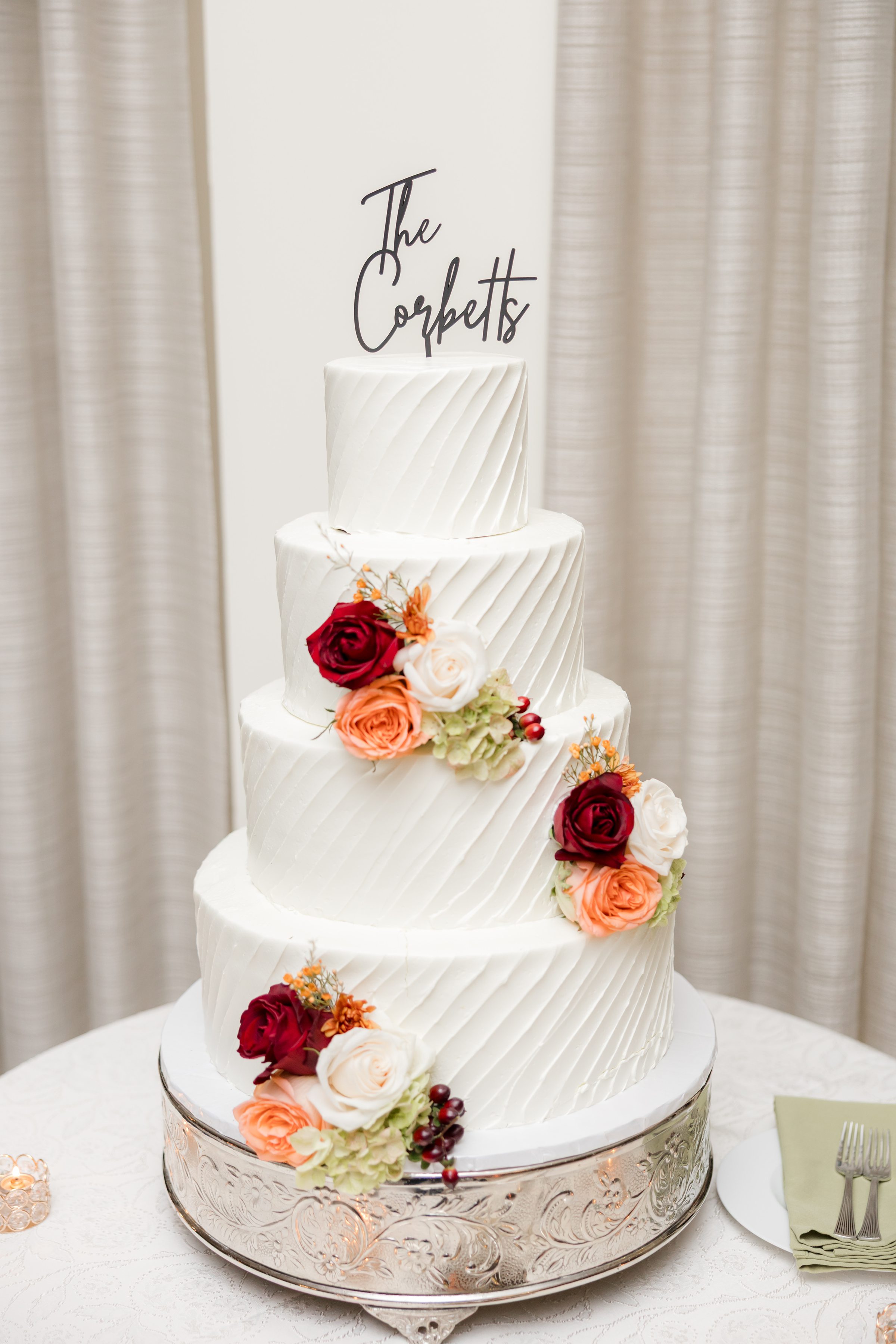 Three-tier white wedding cake with textured icing and floral accents in red, orange, and cream, topped with a “The Corbetts” cake topper in front of soft neutral drapery.
