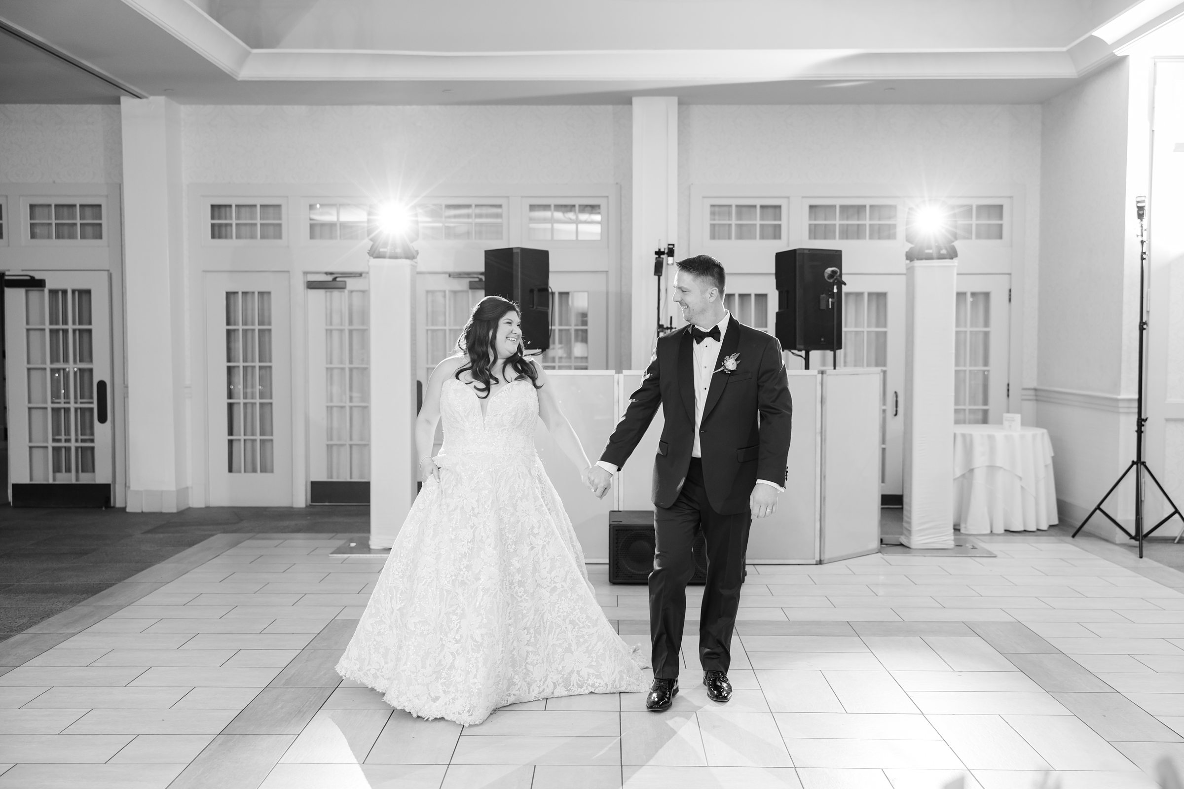 Bride and groom hold hands and smile at each other while walking across the dance floor during their first dance in a bright reception hall