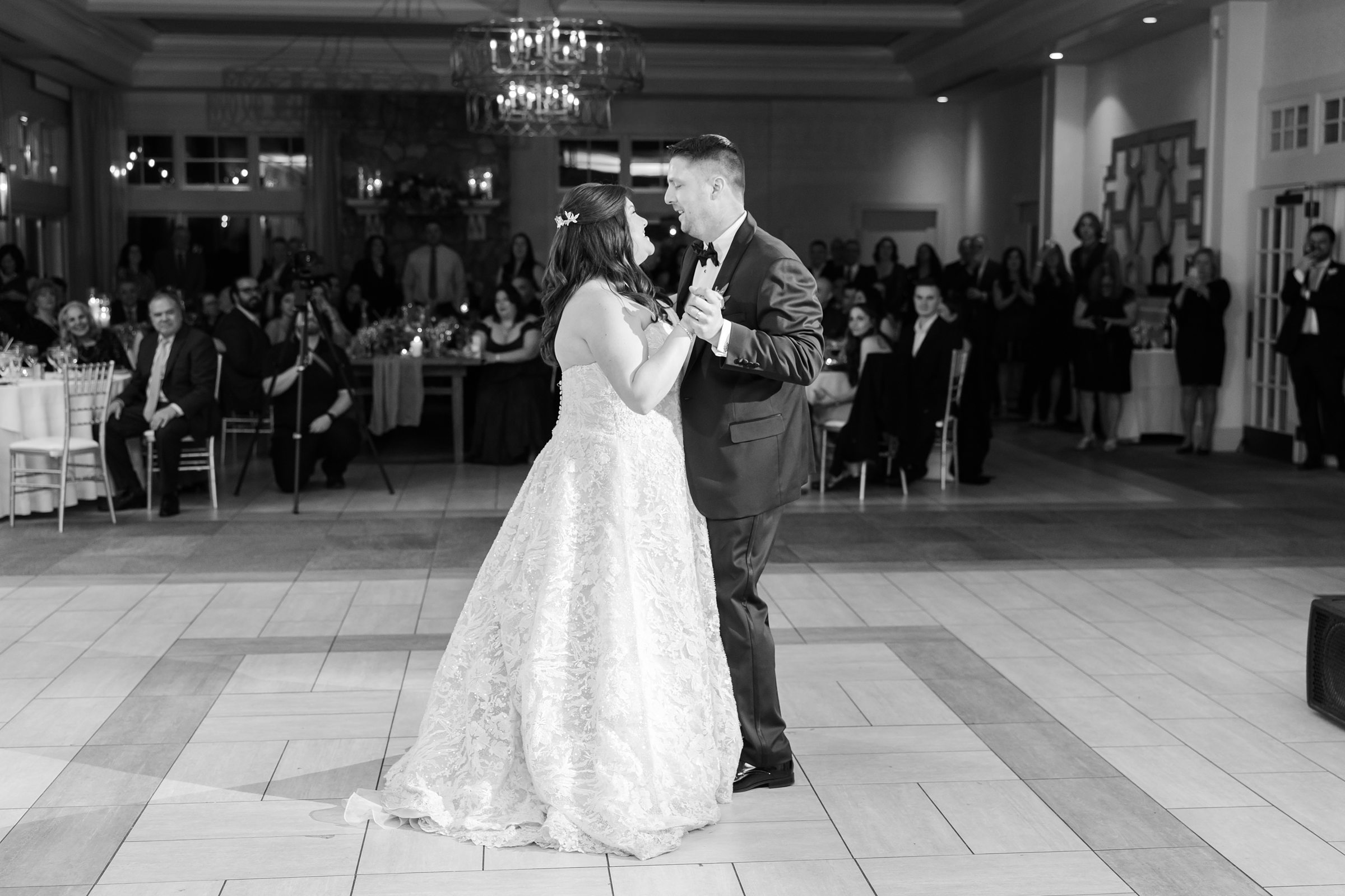 Bride and groom sharing their first dance in the center of a reception hall, surrounded by seated and standing guests watching