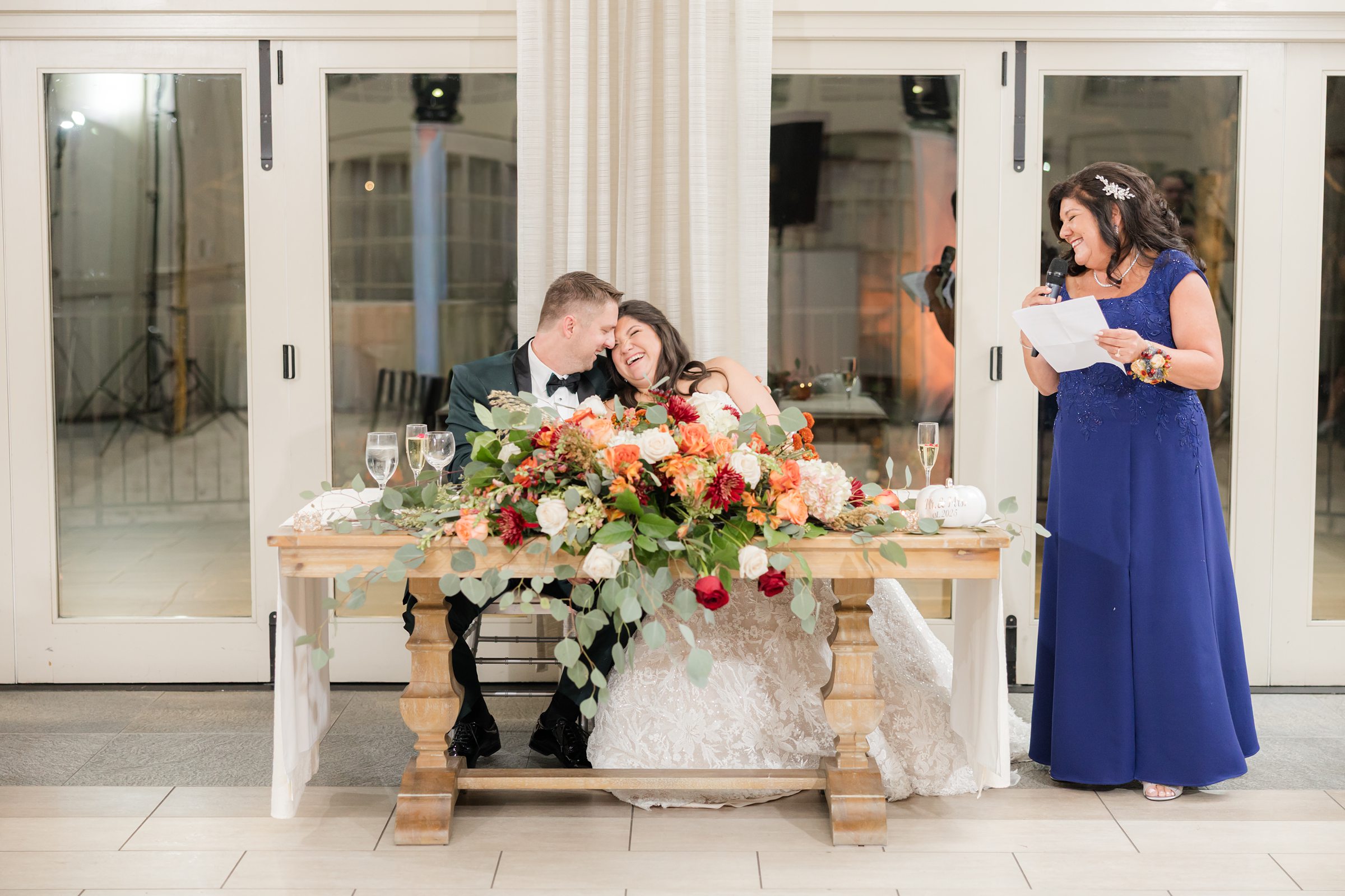 Bride and groom sit close together at their sweetheart table, leaning in and laughing softly as they share a tender moment, while a woman stands beside them giving a heartfelt speech