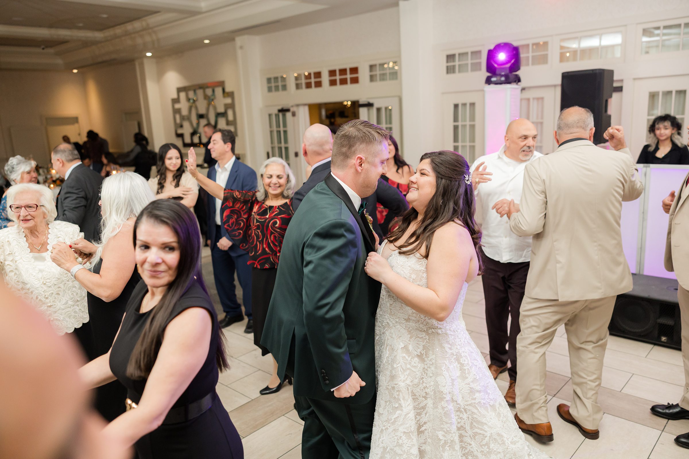 Bride and groom smiles at each other while dancing among guests in a bright reception space