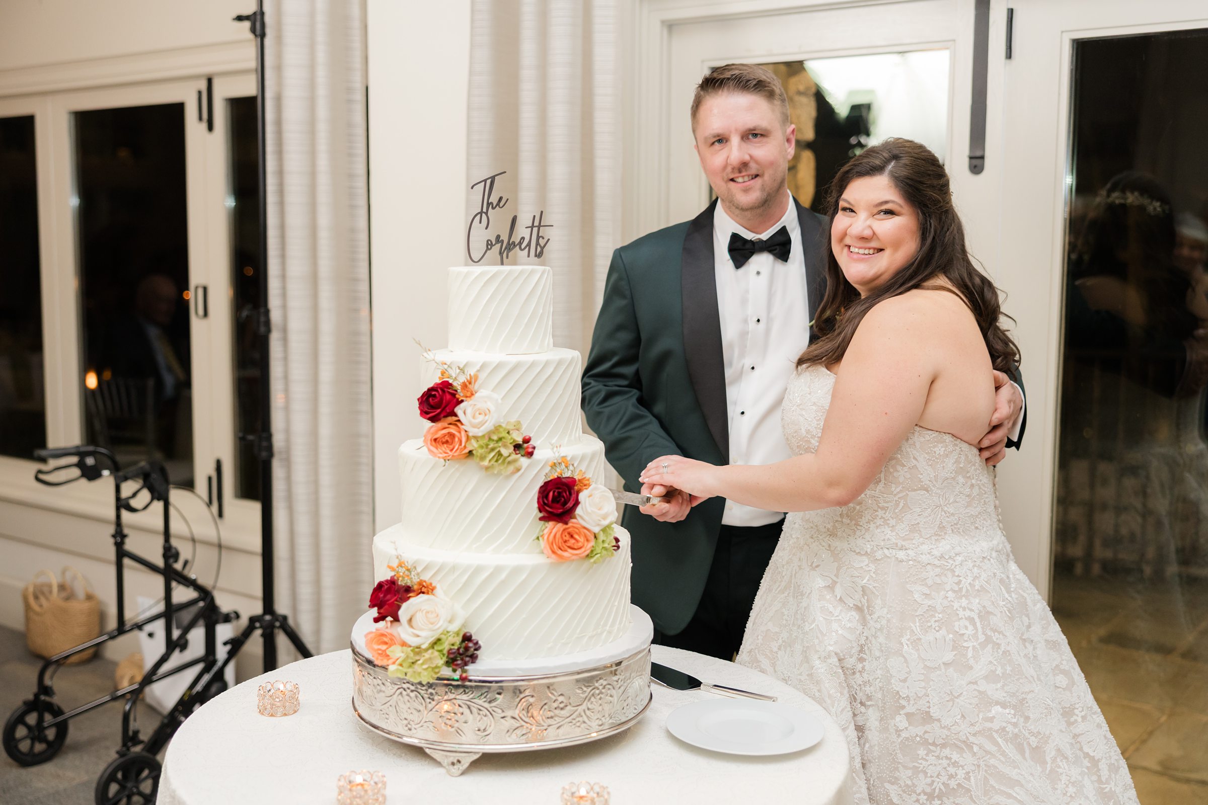 Bride and groom smile while cutting their elegant three-tier wedding cake adorned with red, orange, and cream flowers.