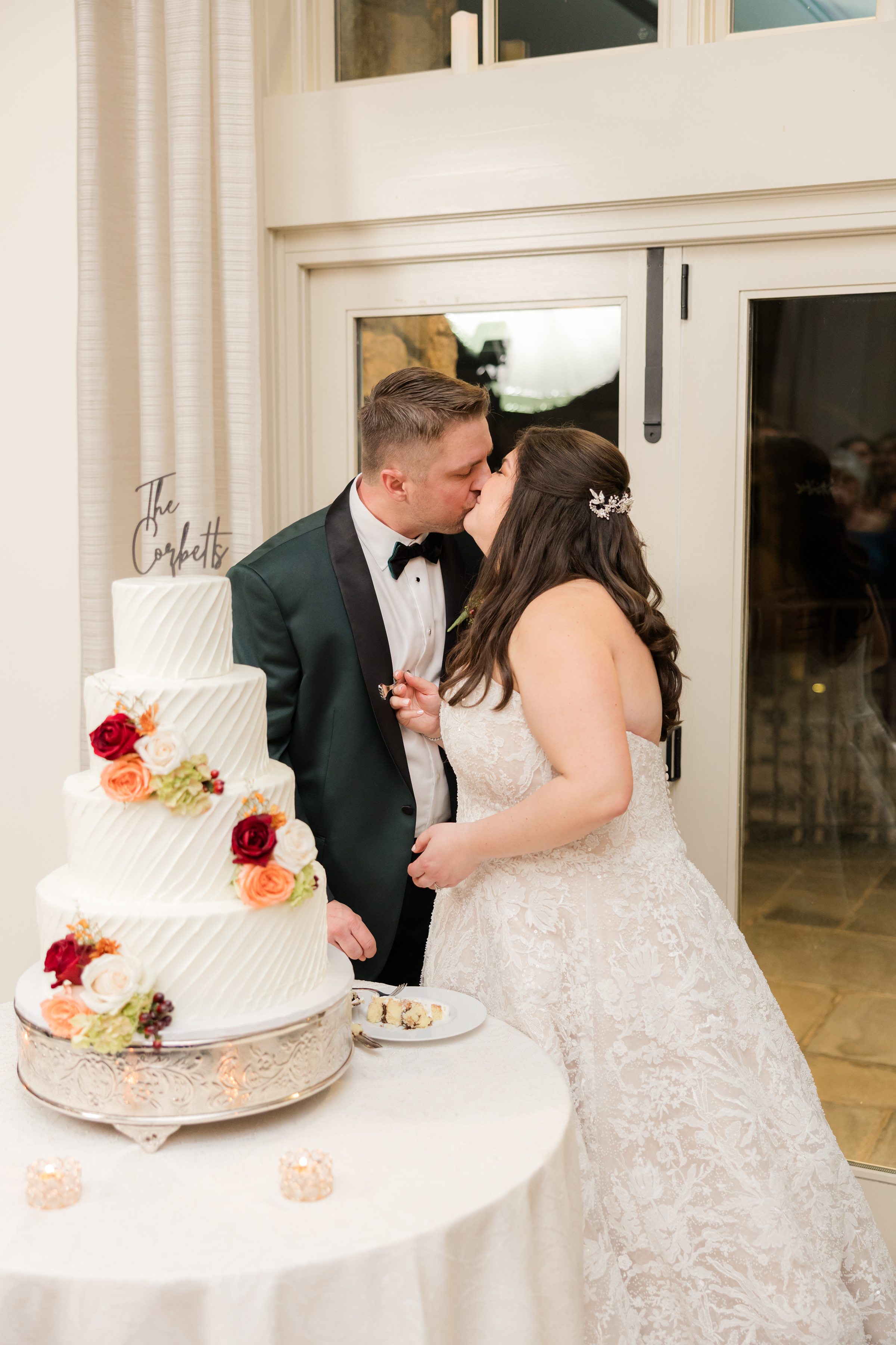 Bride and groom kiss beside a three-tier white wedding cake decorated with red, orange, and cream flowers