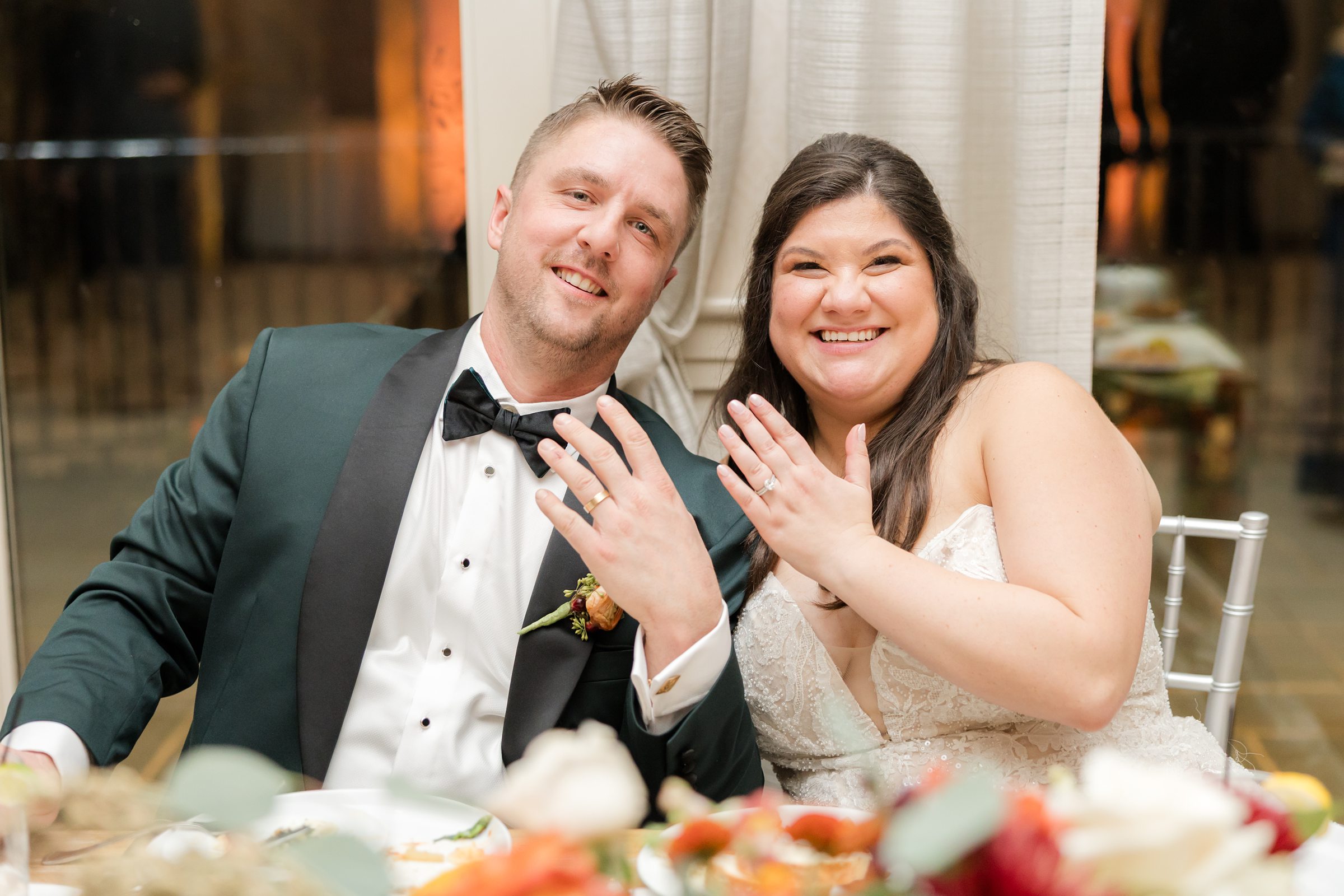 Bride and groom sit together smiling brightly and holding up their hands to show their wedding rings, sharing a joyful and celebratory moment